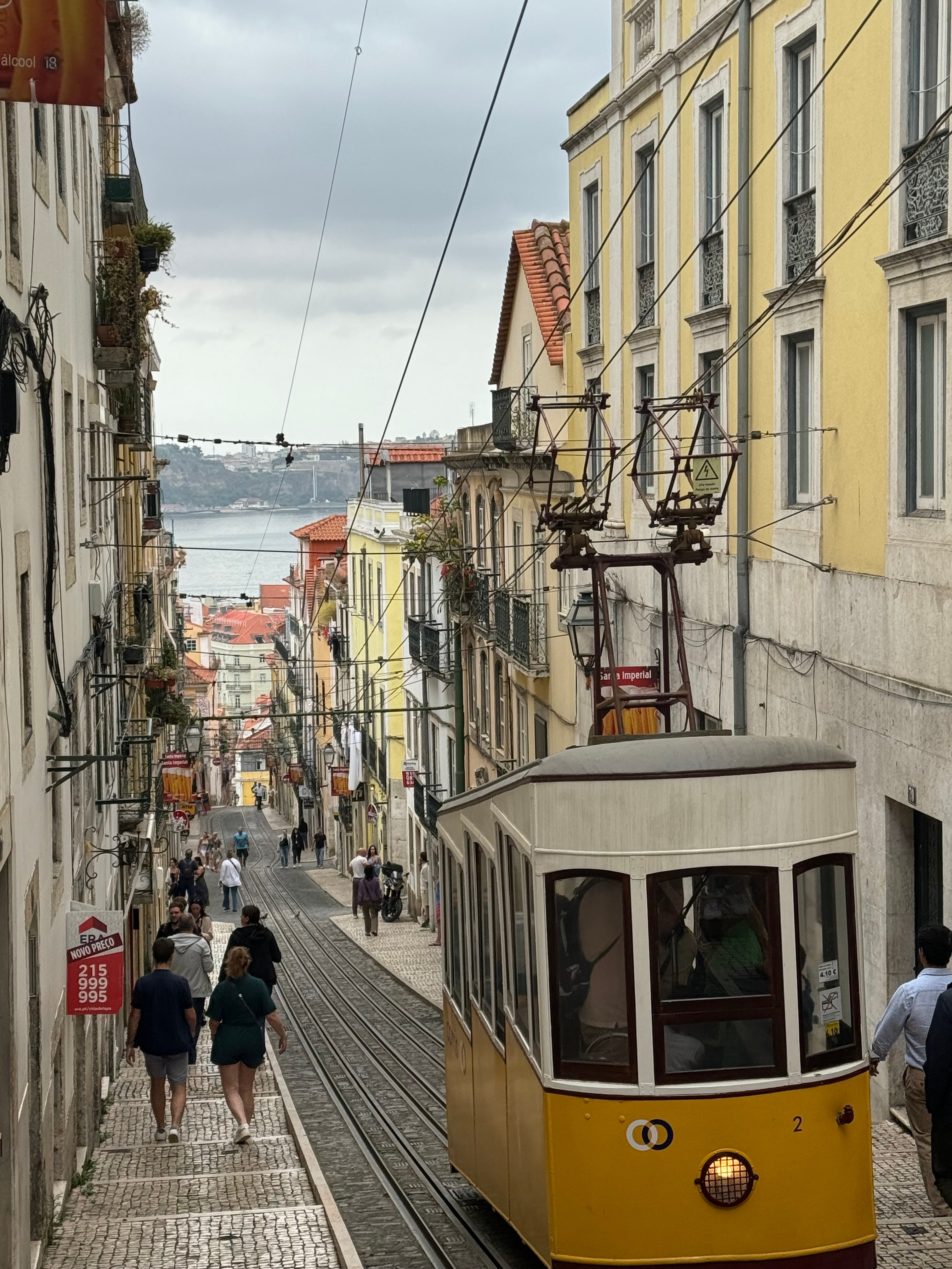 Eléctrico histórico numa rua íngreme em Lisboa, Portugal, com pessoas a caminhar e vistas distantes da água e da arquitectura tradicional