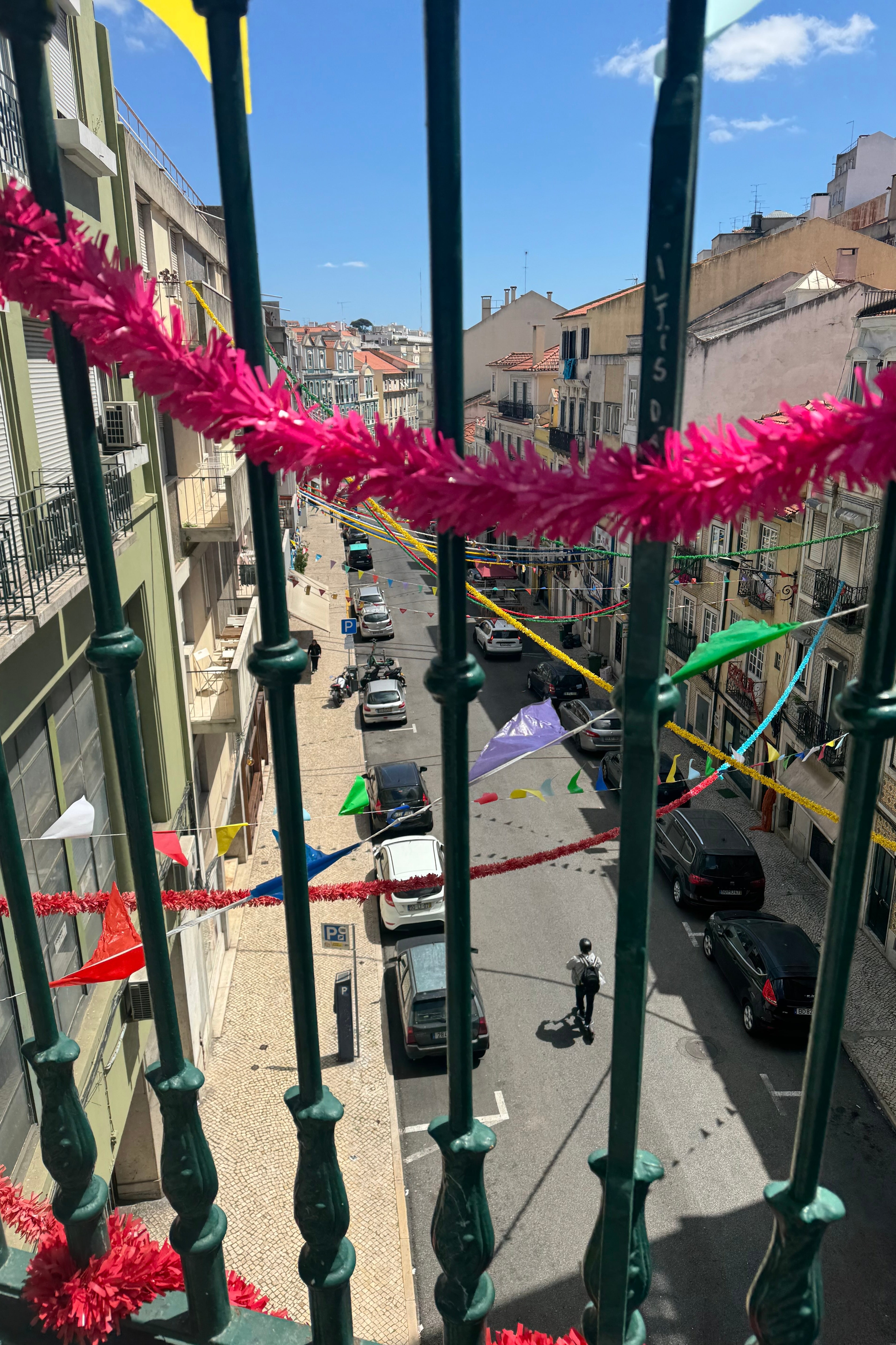 Vista de uma varanda com vista para uma rua movimentada de uma cidade, decorada com faixas e serpentinas coloridas, sugerindo um clima festivo
