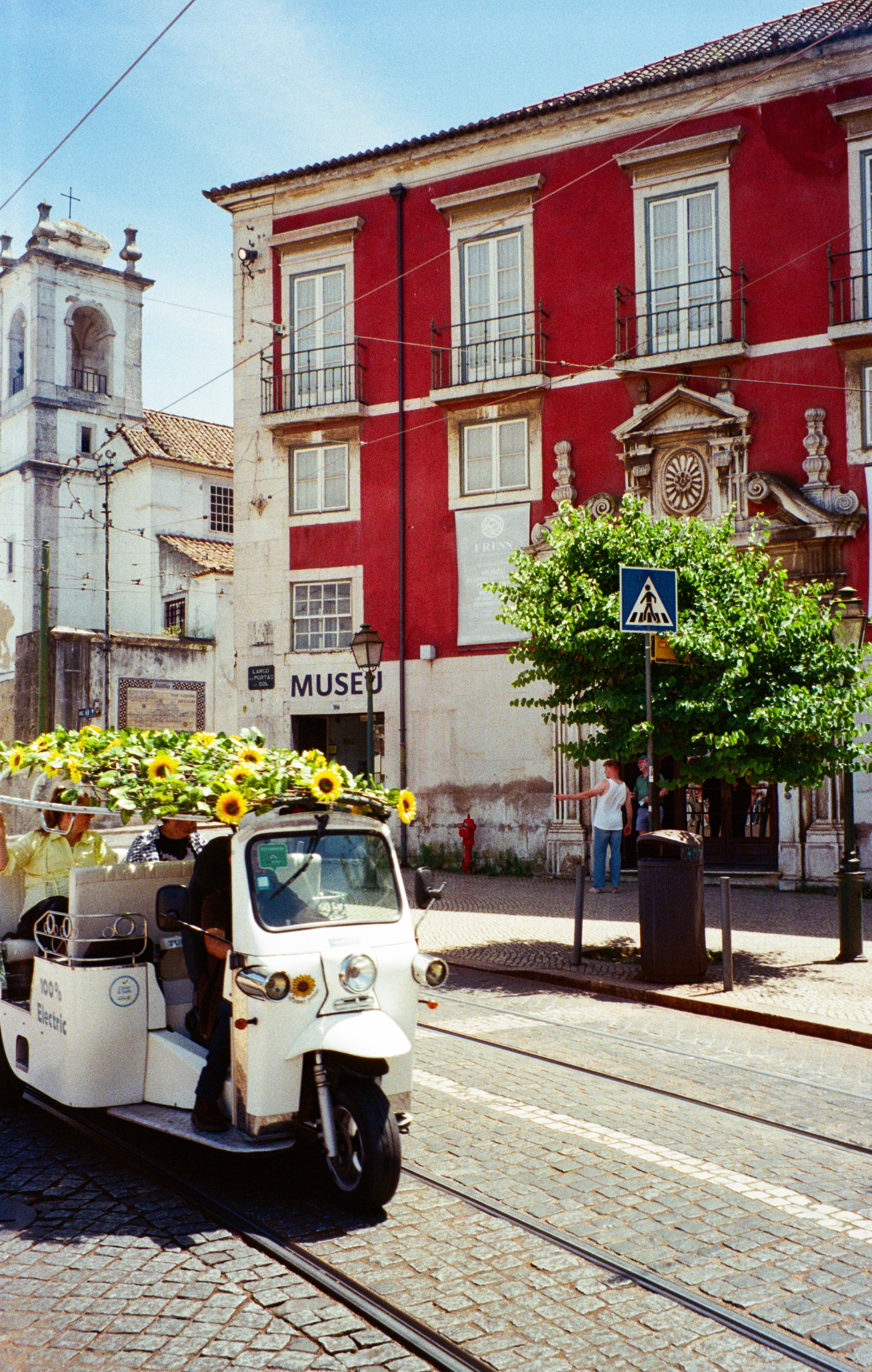 Um tuk-tuk elétrico branco com girassóis no topo passa por um edifício histórico em uma rua de paralelepípedos em uma cidade europeia