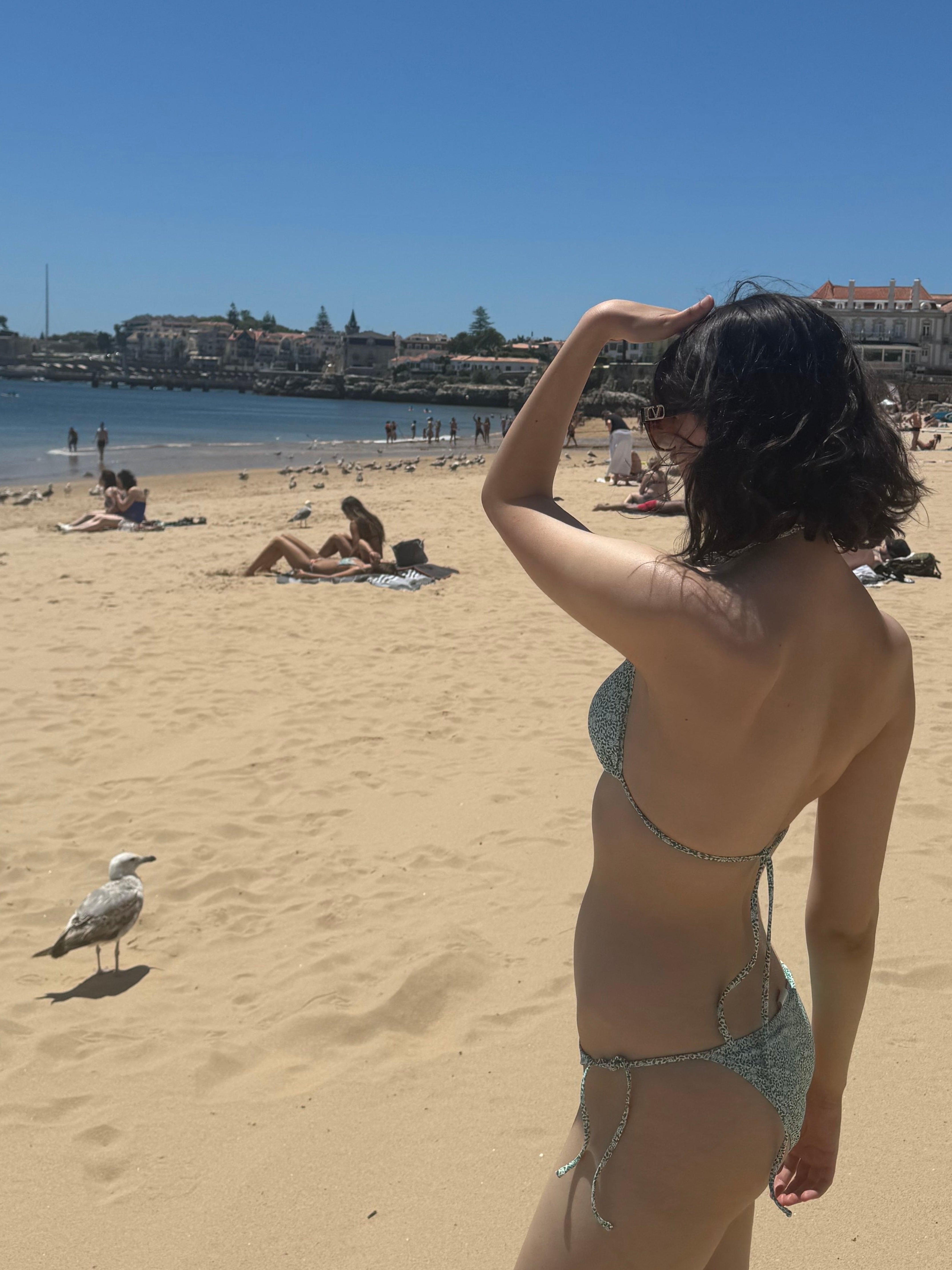 Pessoa de biquíni fica em uma praia ensolarada, protegendo os olhos enquanto olha para longe; gaivota por perto, pessoas ao fundo