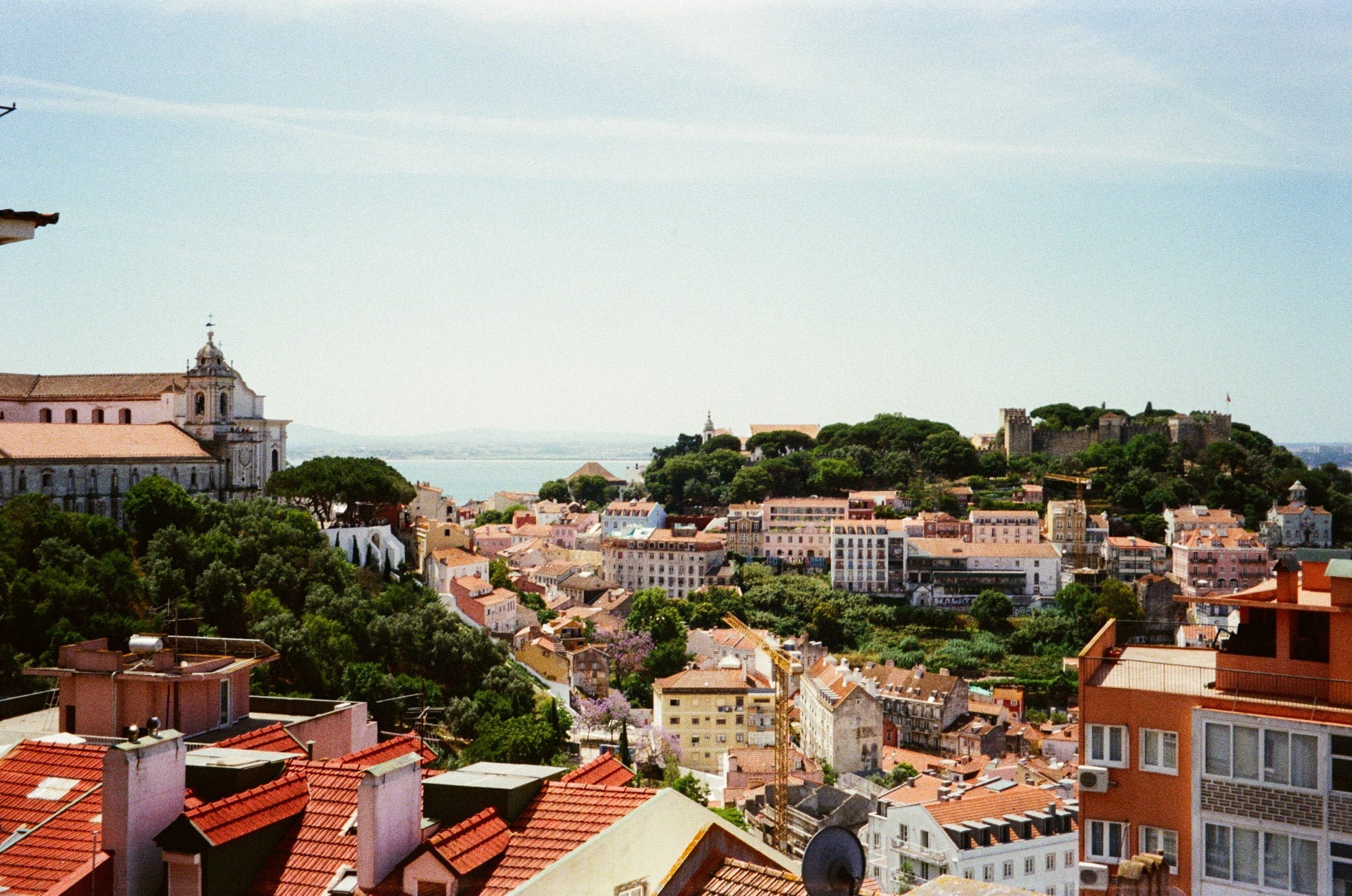 Vista panorâmica dos telhados de Lisboa com o Castelo de São Jorge e a vegetação circundante ao fundo sob um céu claro