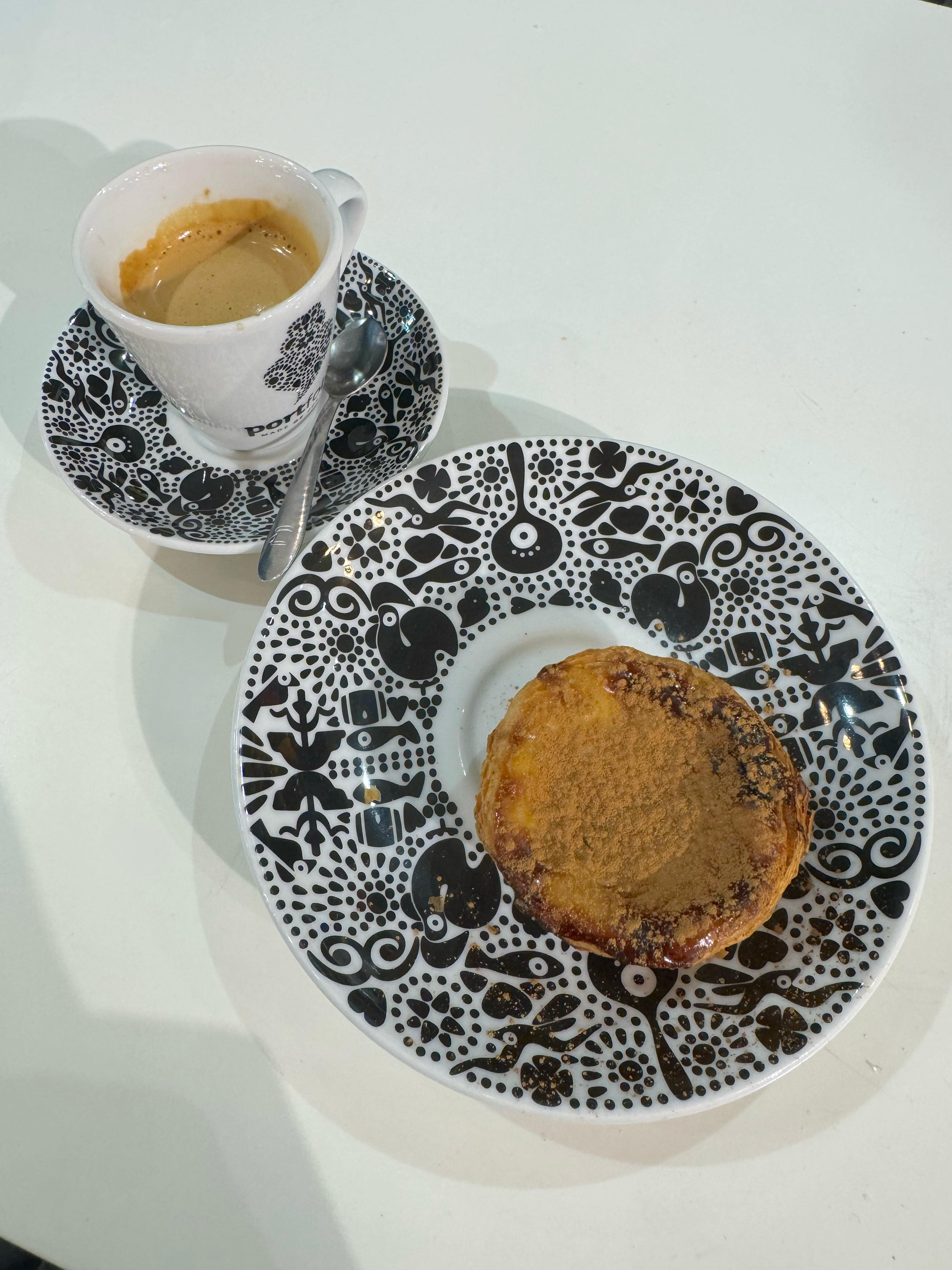 Uma xícara de café expresso com uma colher em um pires estampado ao lado de um doce em um prato correspondente, colocado sobre uma mesa branca