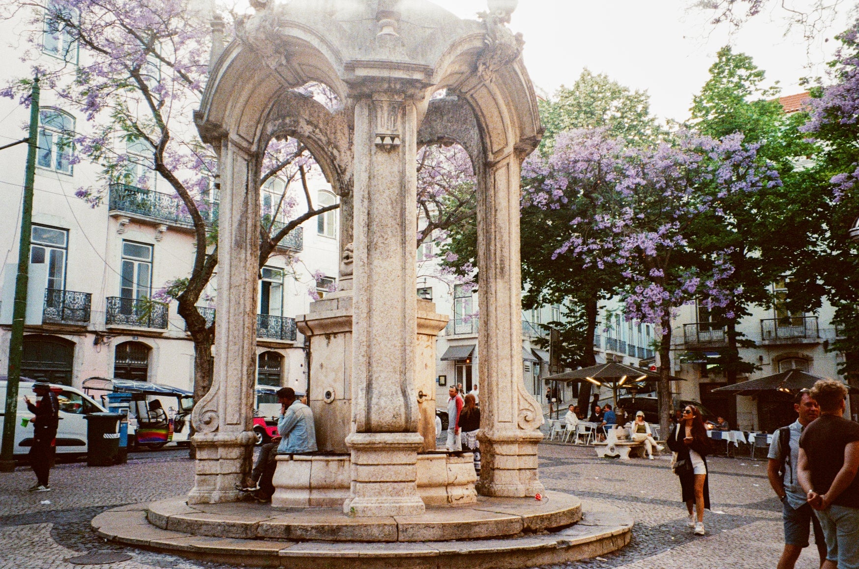 Pessoas sentadas perto de uma fonte de pedra ornamentada em uma praça vibrante com árvores floridas ao fundo. Vibração arquitetônica europeia