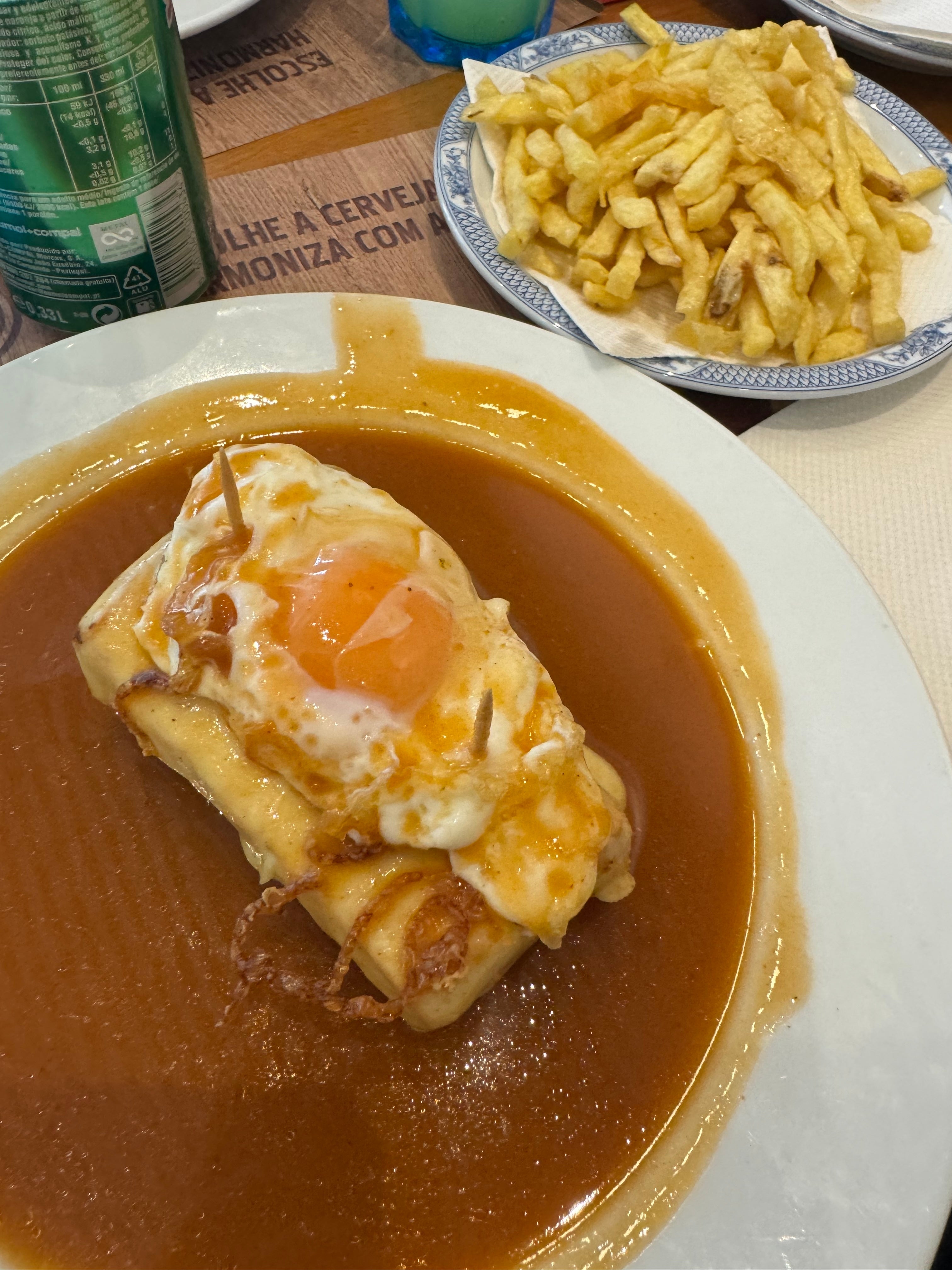 Prato com sanduíche de francesinha com ovo por cima, rodeado de molho e acompanhamento de batata frita