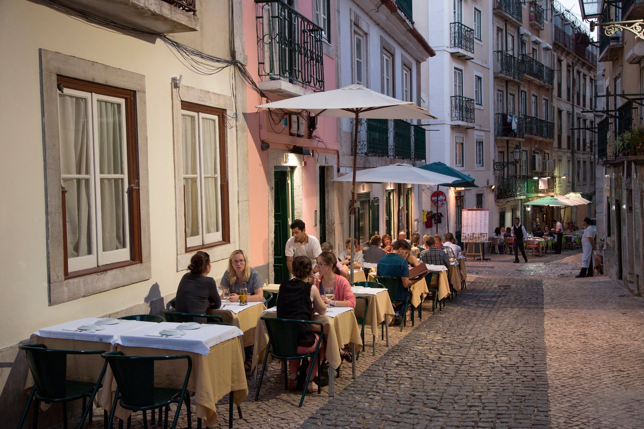 Pessoas jantando em mesas ao ar livre ao longo de uma rua estreita de paralelepípedos em uma cidade europeia, com prédios e guarda-sóis criando um ambiente aconchegante