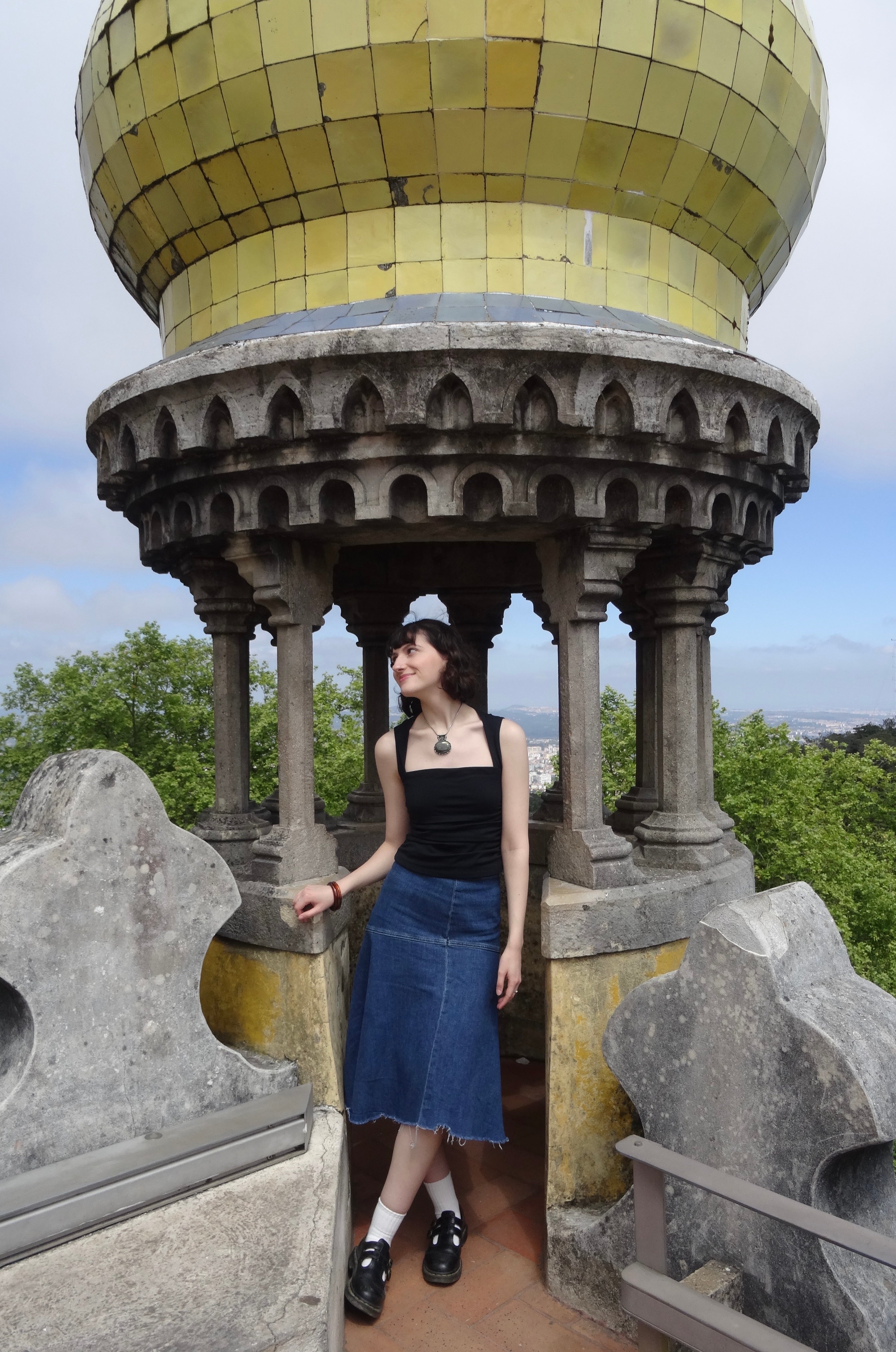 Pessoa com roupa casual fica em um terraço de pedra de um edifício histórico com colunas ornamentadas, com vista para uma paisagem verdejante