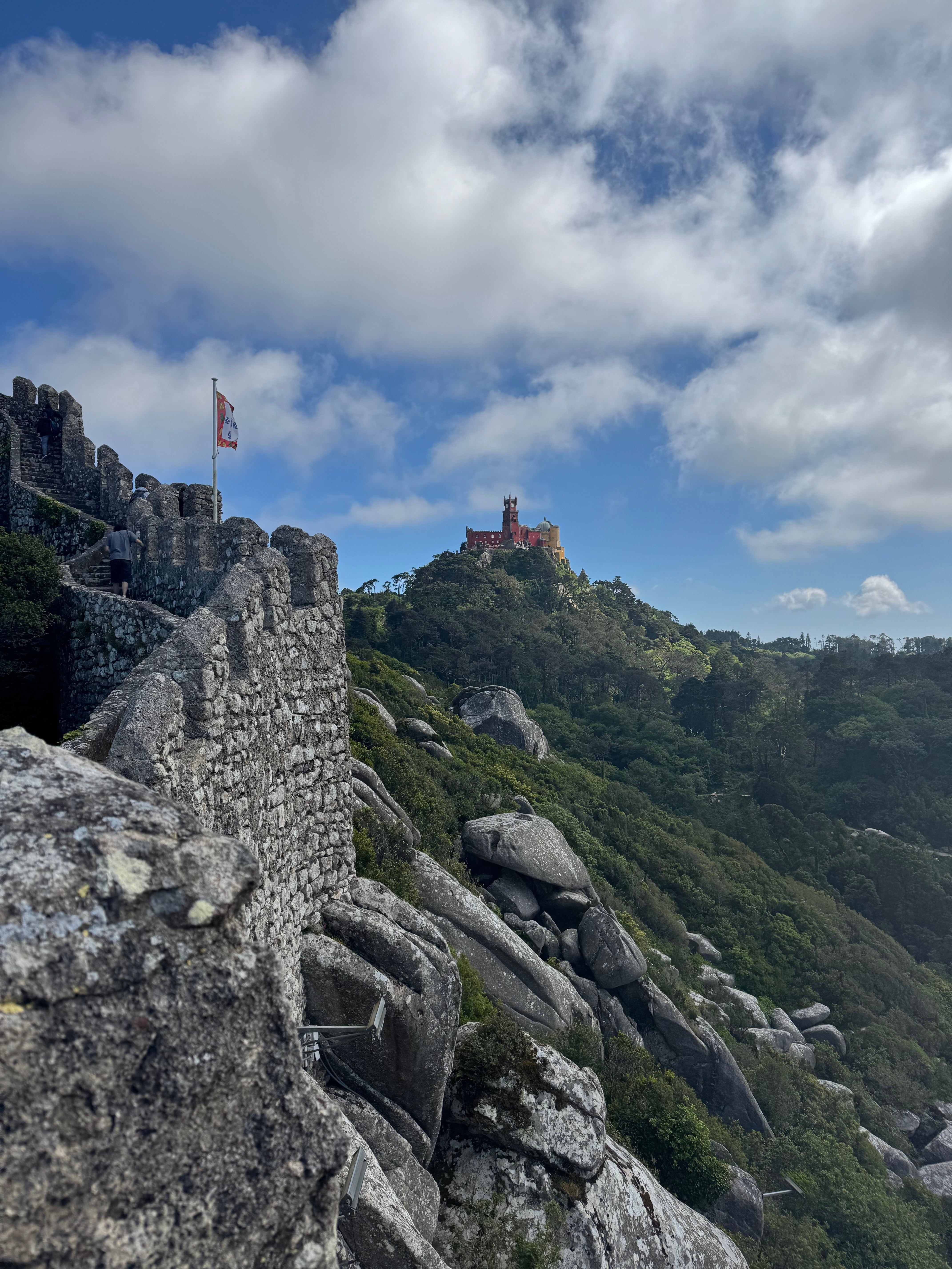 Um castelo fica em uma colina rochosa exuberante sob um céu parcialmente nublado, com antigas paredes de pedra em primeiro plano