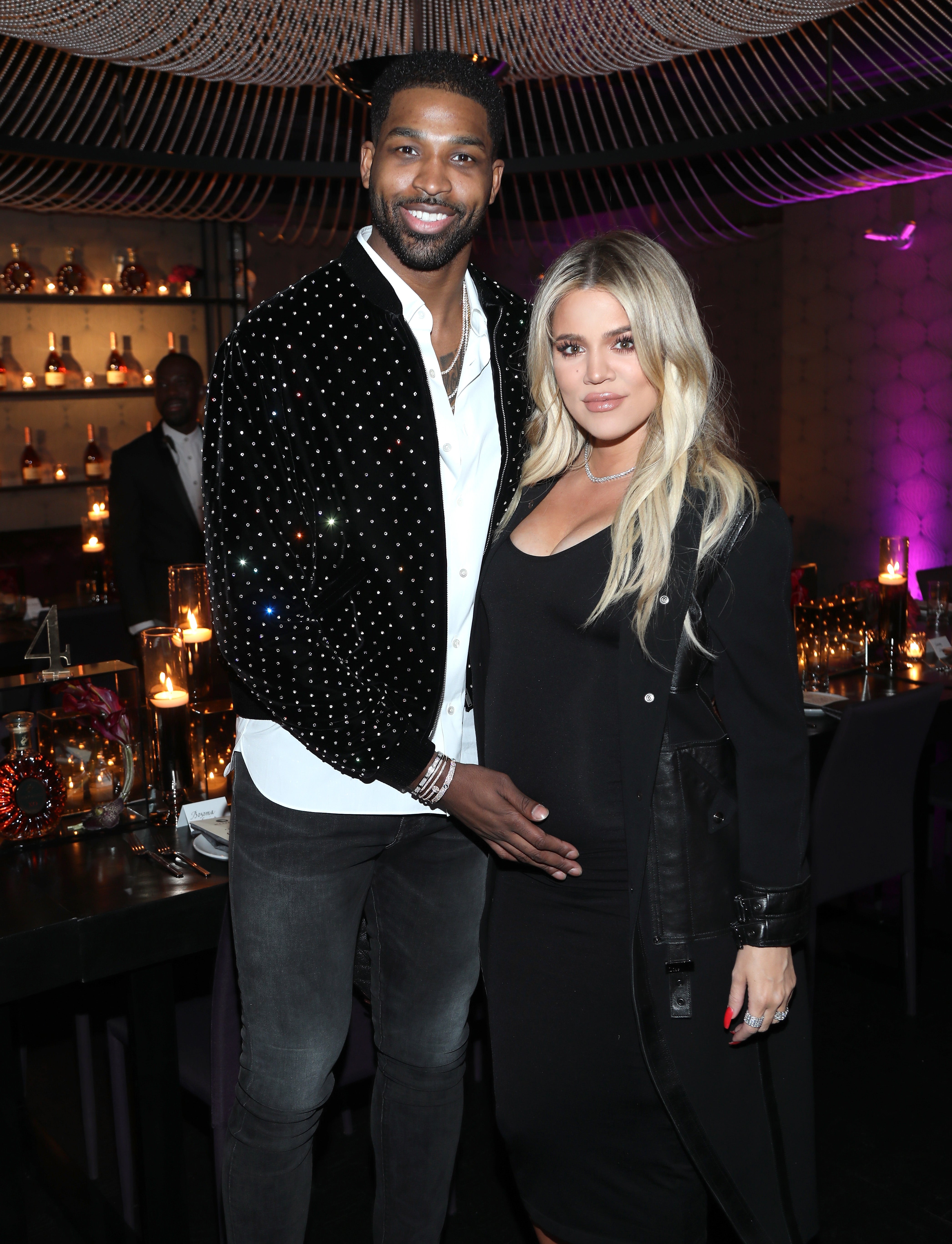 Tristan Thompson wears a bedazzled jacket and poses with his hand on Khloe Kardashian's stomach. Khloe Kardashian wears a black dress and black jacket