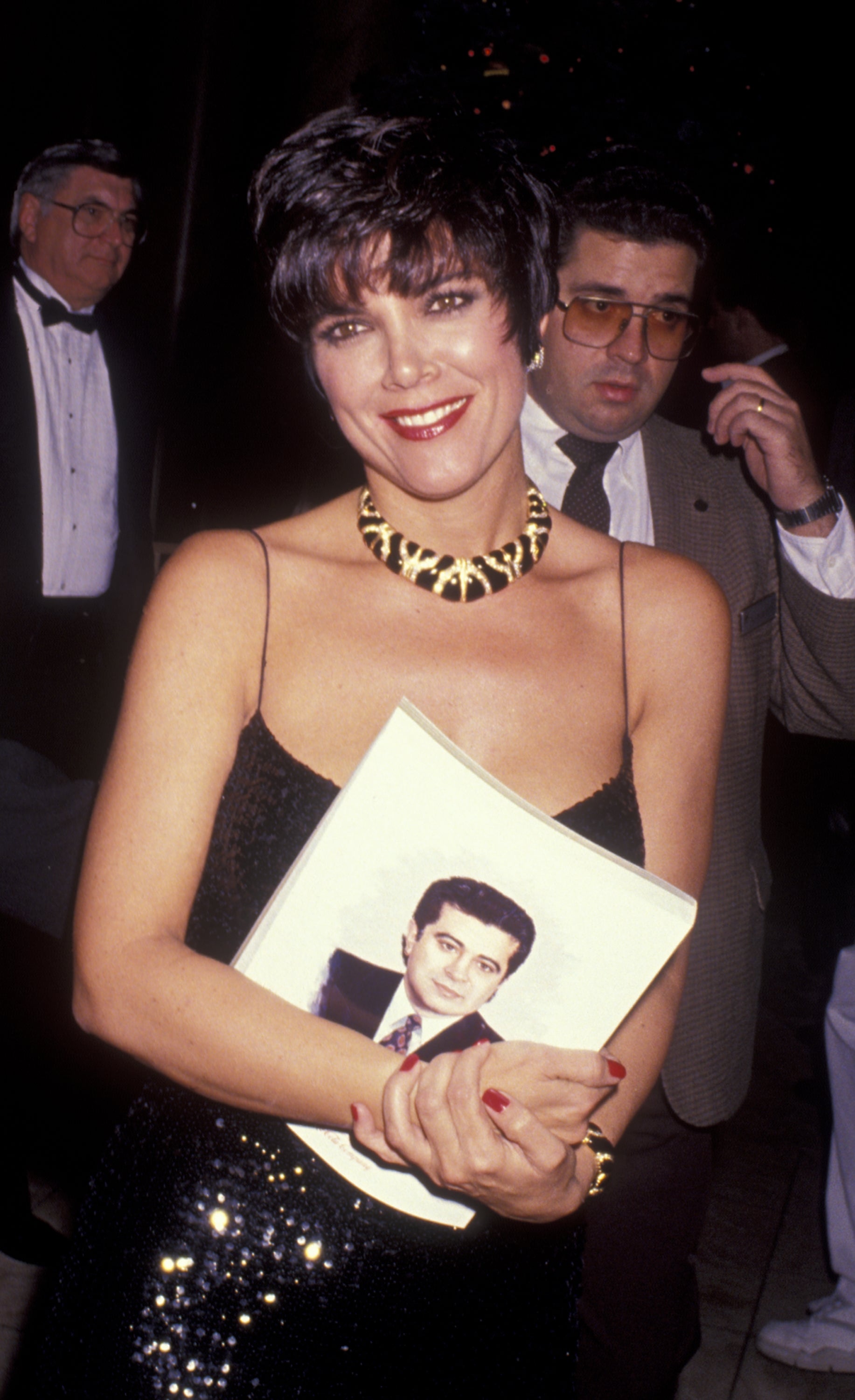 Kris Jenner smiles while holding a portrait of a man wearing a black, sequined gown