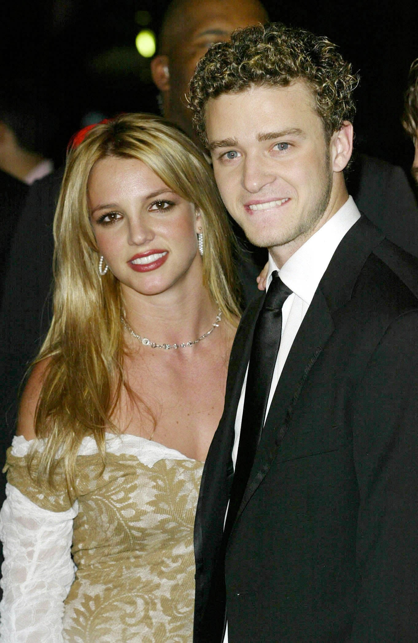 Britney Spears wears a gold and white patterned dress while posing beside Justin Timberlake who wears a black suit and bites his lip