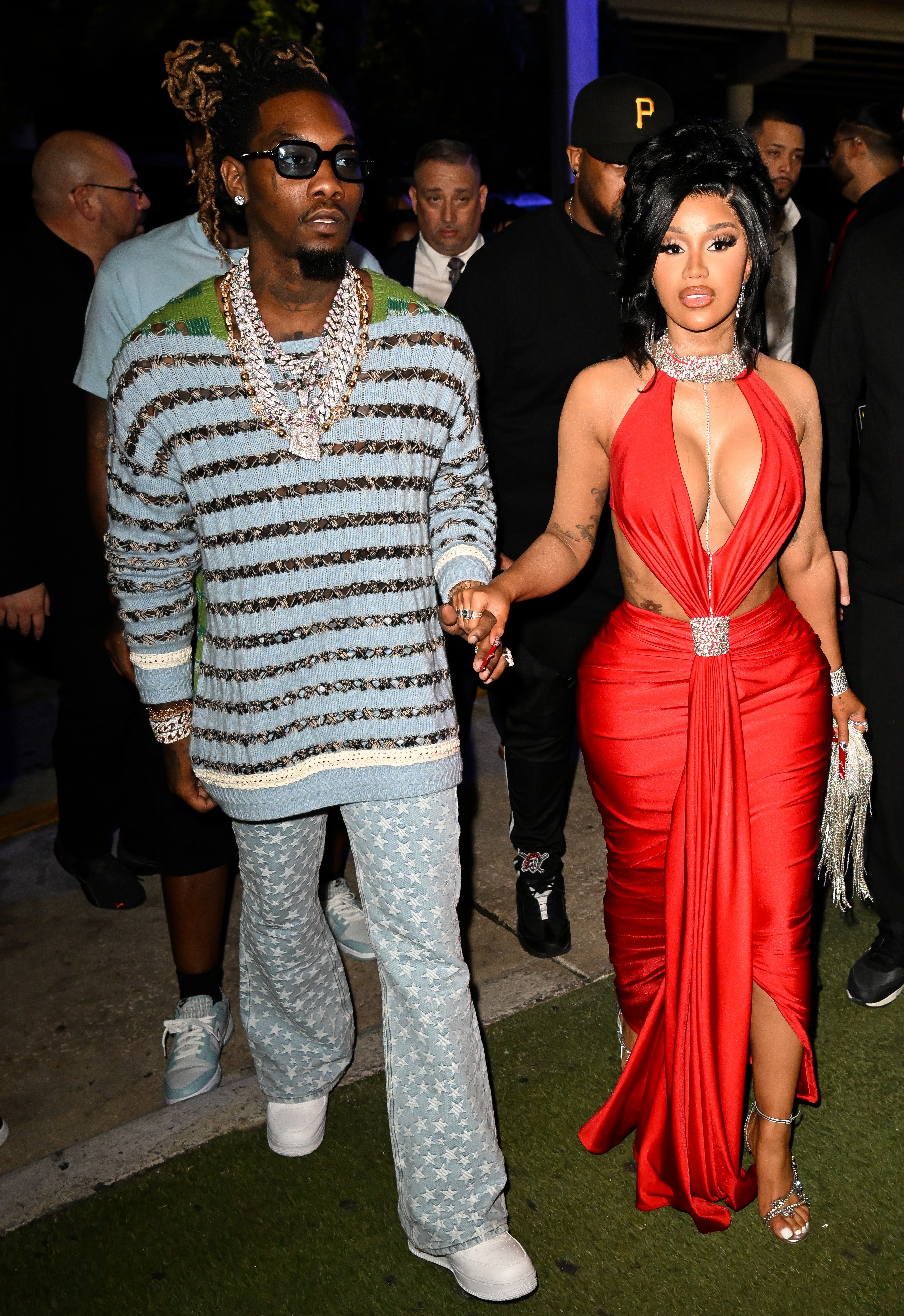 Offset and Cardi B hold hands as they walk together. Offset wears a sky blue sweater with star-patterned jeans and Cardi B wears a red dress