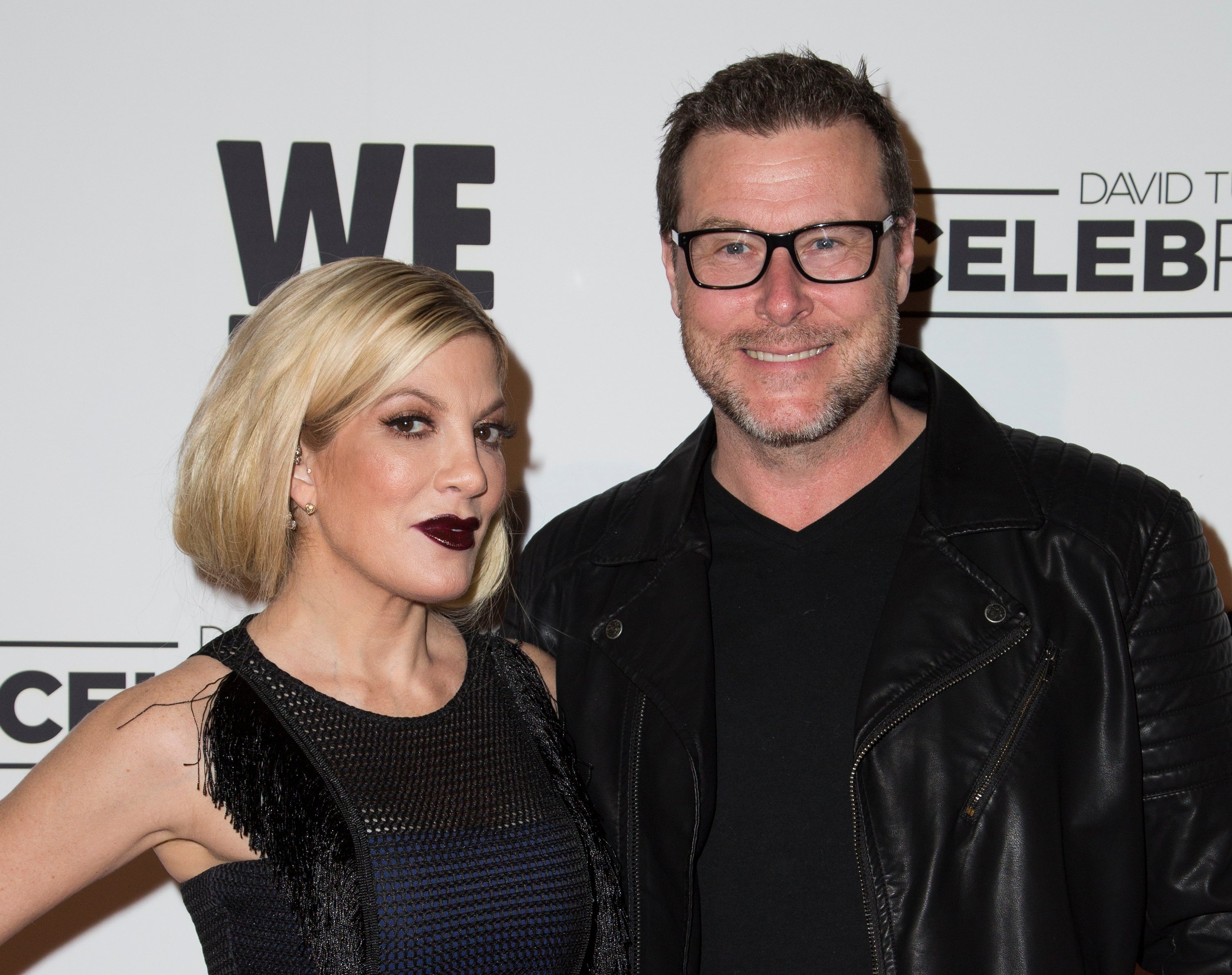 Tori Spelling wears dark lipstick and a black dress while posing with Dean McDermott who wears a black shirt and jacket