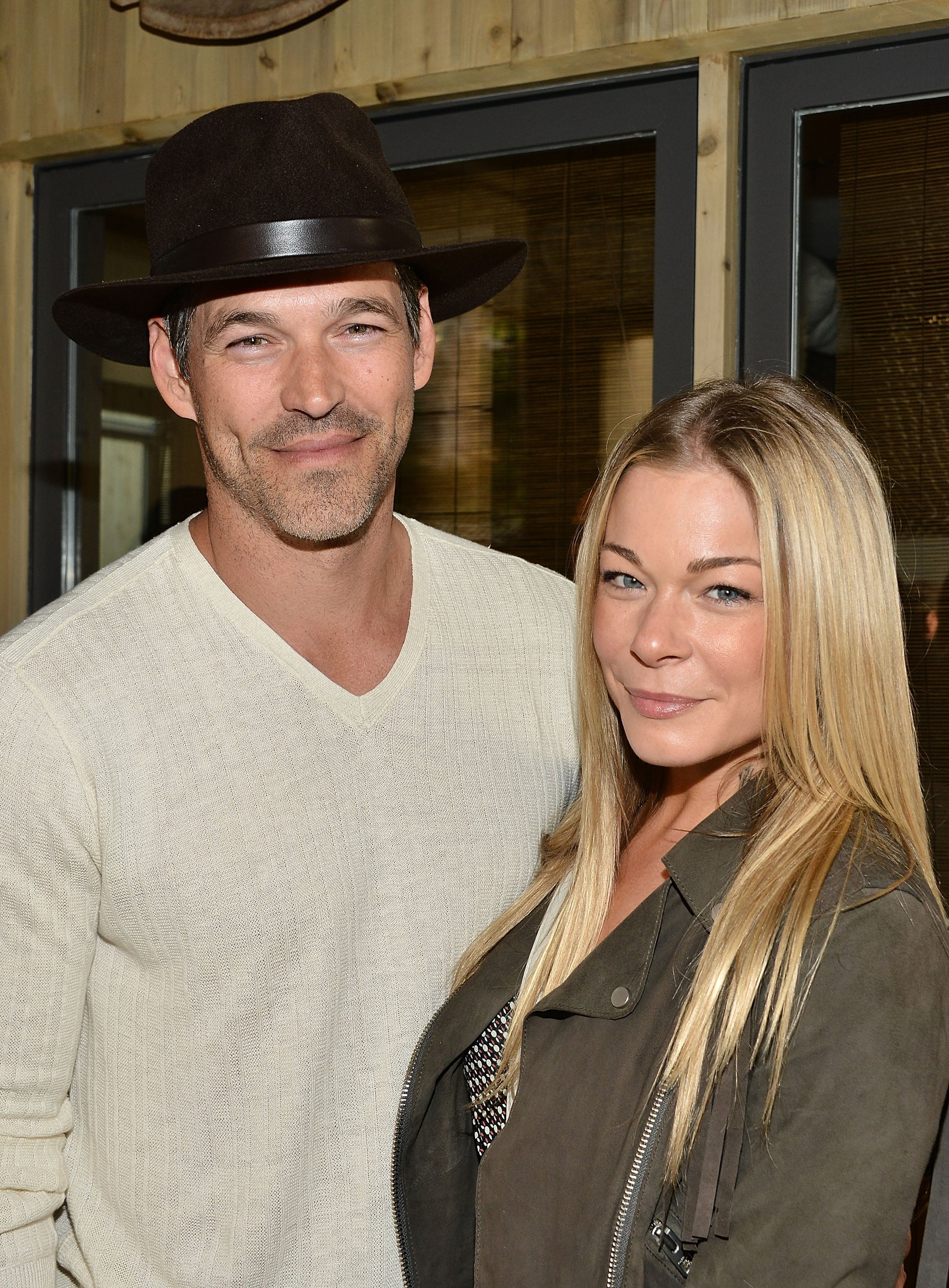 Eddie Cibrian wears a large hat and white sweater while posing beside LeAnn Rimes who wears an olive green jacket