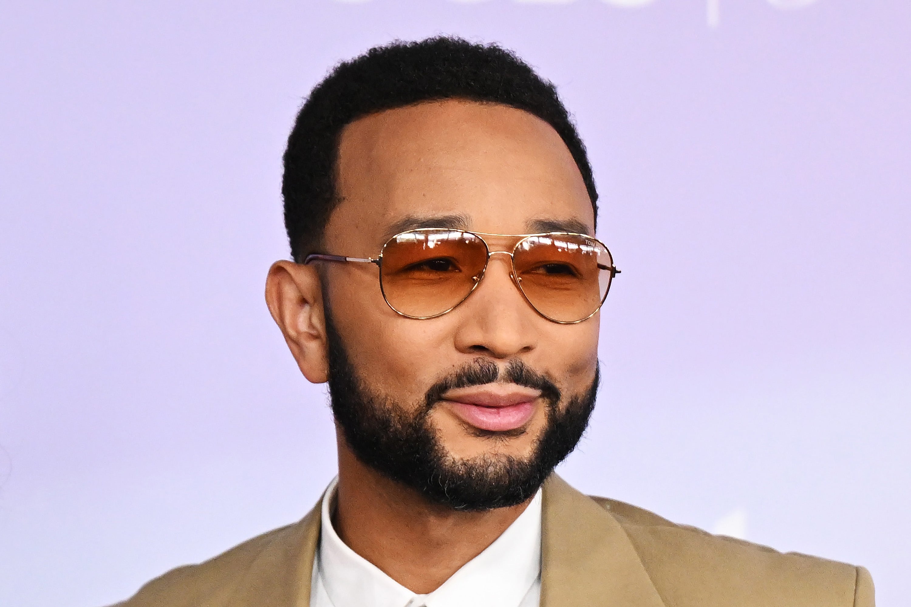 John Legend poses in front of a lavender background wearing brown aviator sunglasses