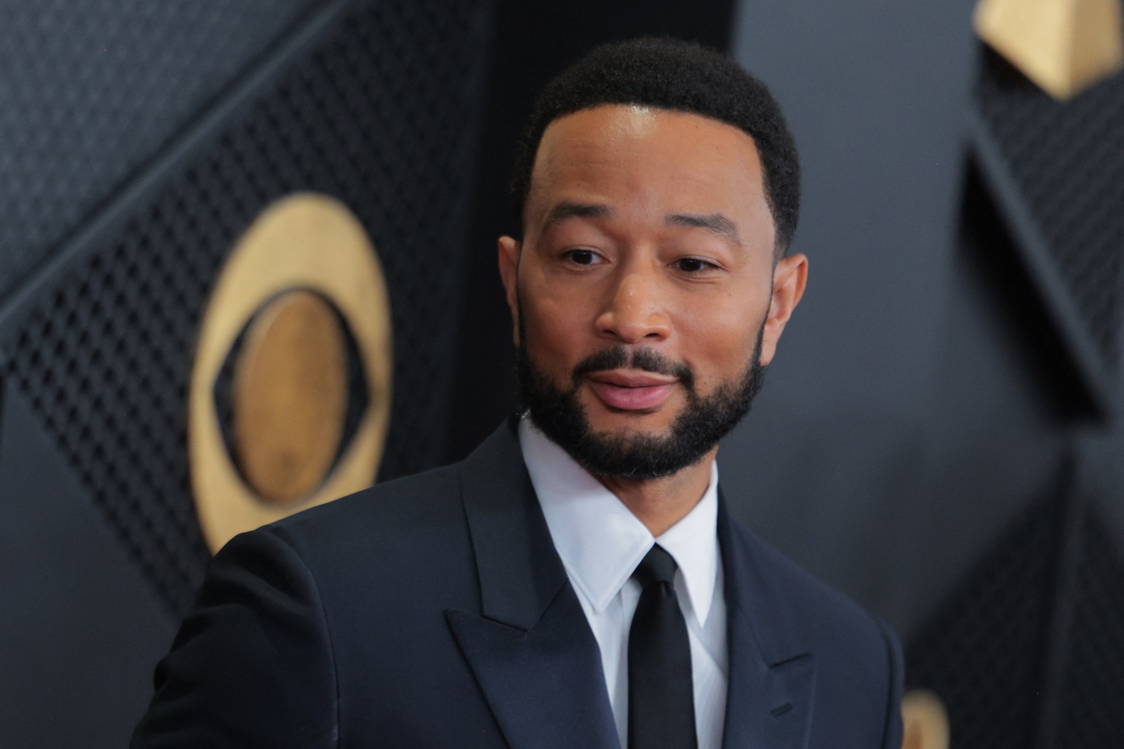 John Legend poses at an event wearing a black suit and tie