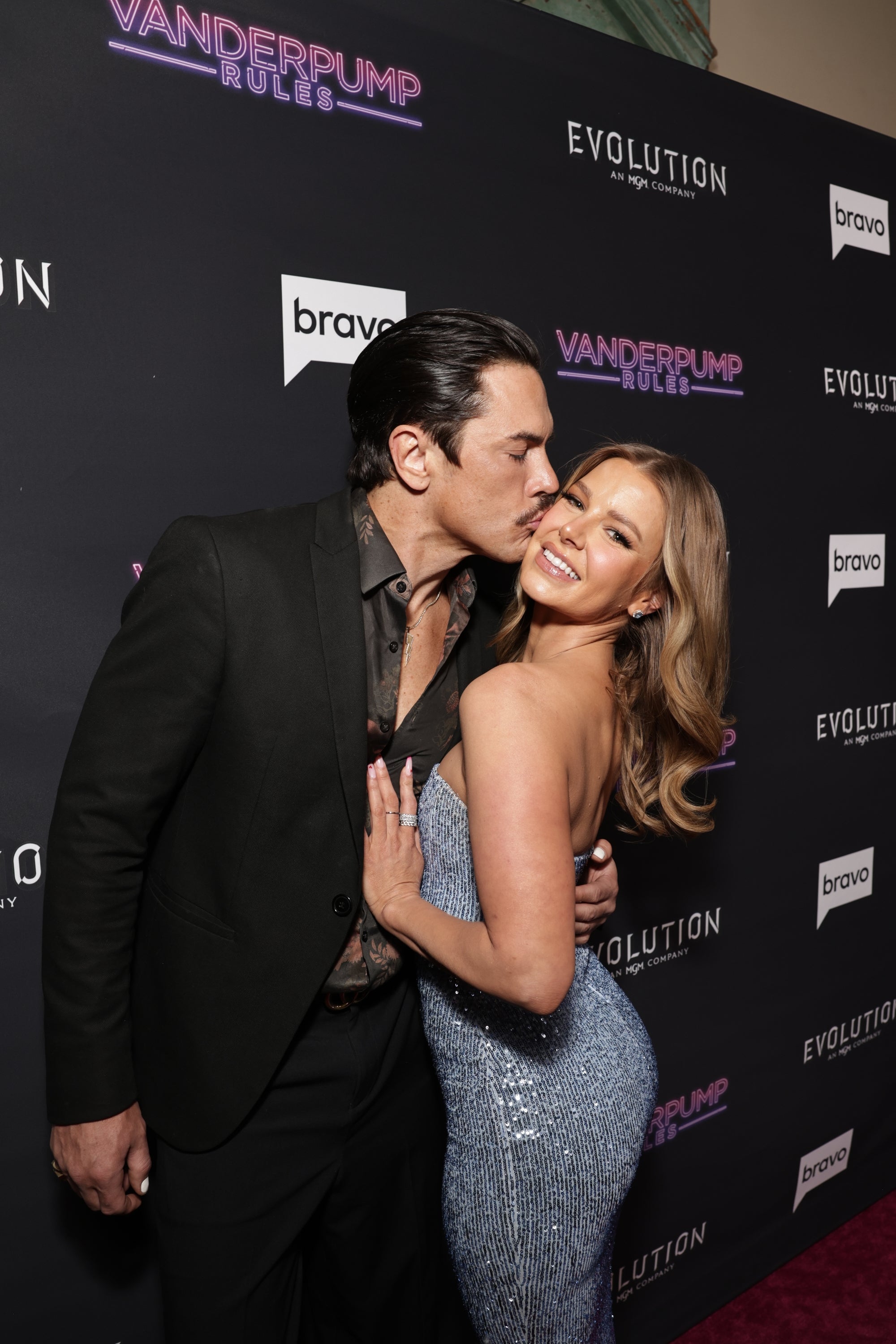 Tom Sandoval kisses Ariana Madix's cheek on a red carpet. Ariana Madix wears a silver, sequined gown