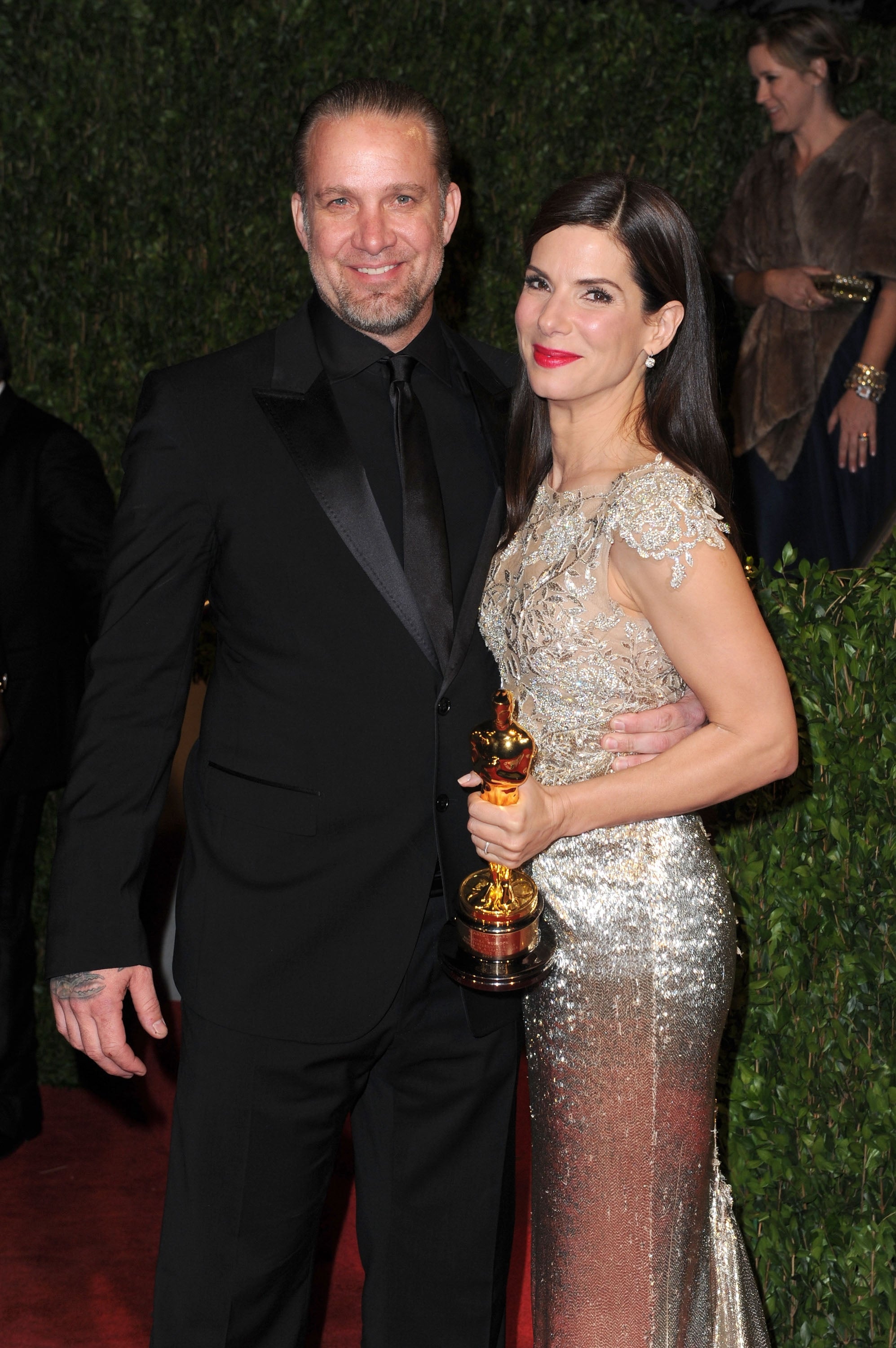 Jesse James wears a black suit and poses with Sandra Bullock. Sandra Bullock wears a sparkling gown and holds an Academy Award in her hand