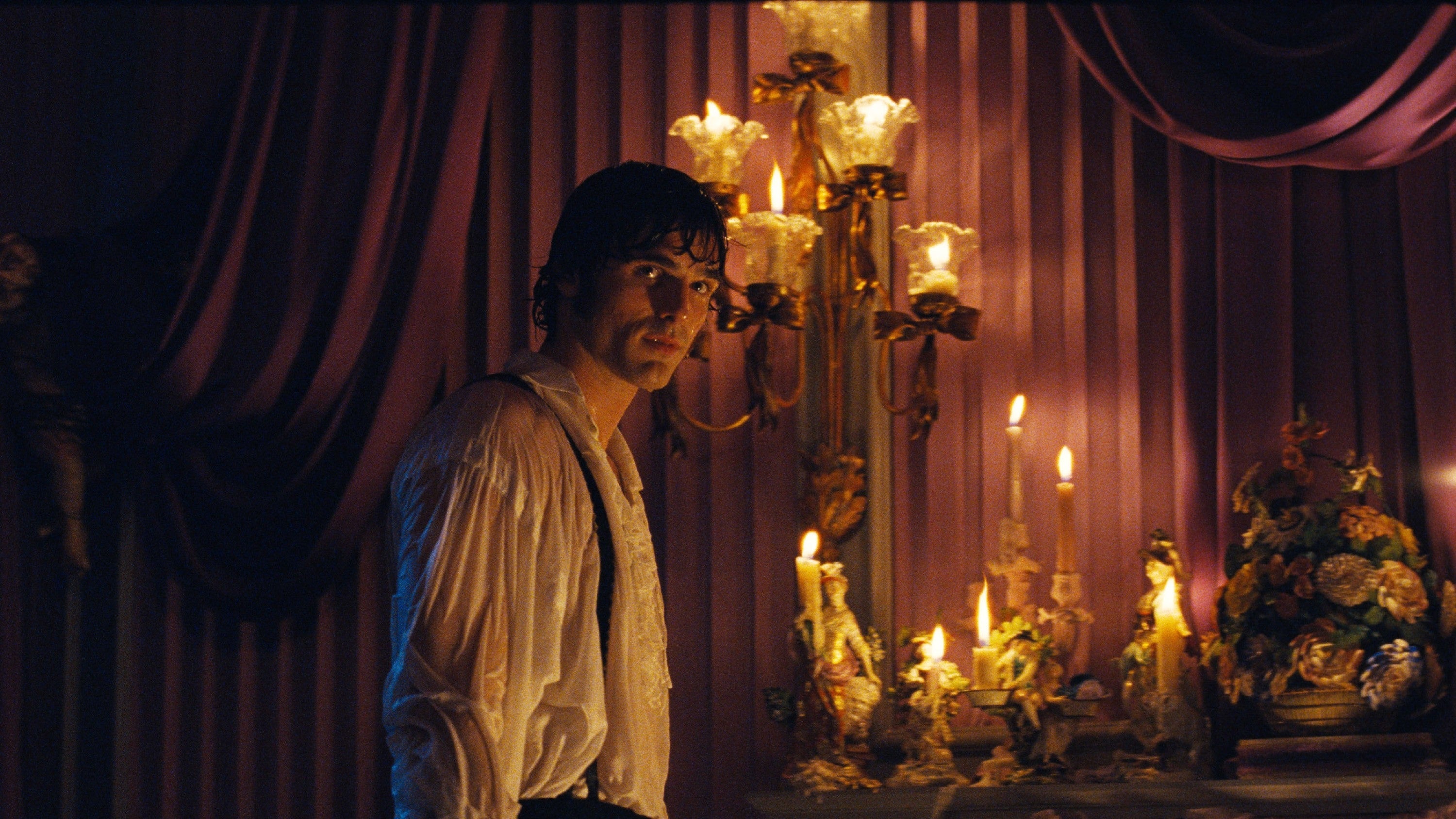 Jacob Elordi in period attire stands beside ornate candelabra and drapes, evoking a historical or theatrical setting