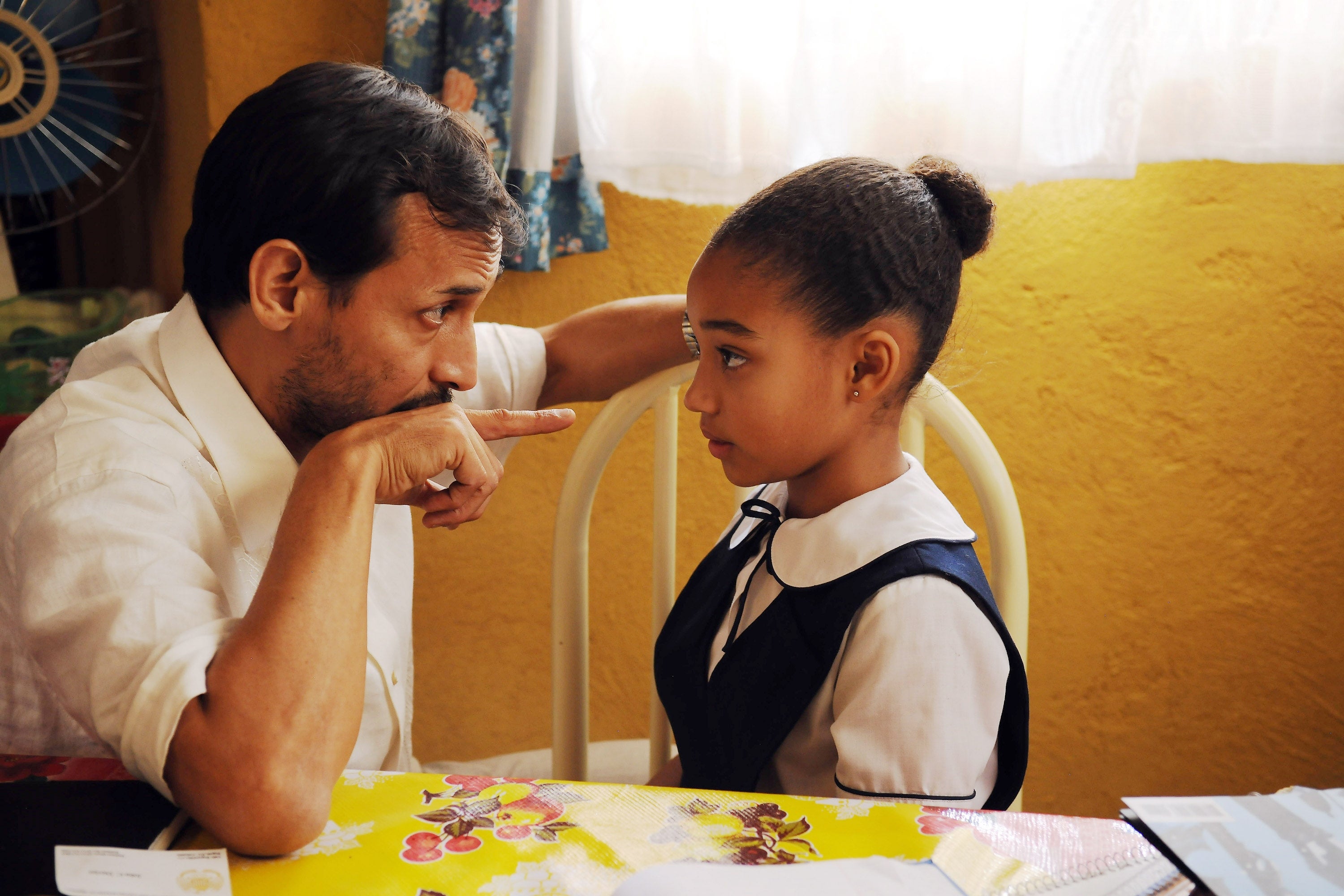 Jesse Borrego sitting at a table, intently talking to young Amandla Stenberg in a school uniform, who is listening seriously