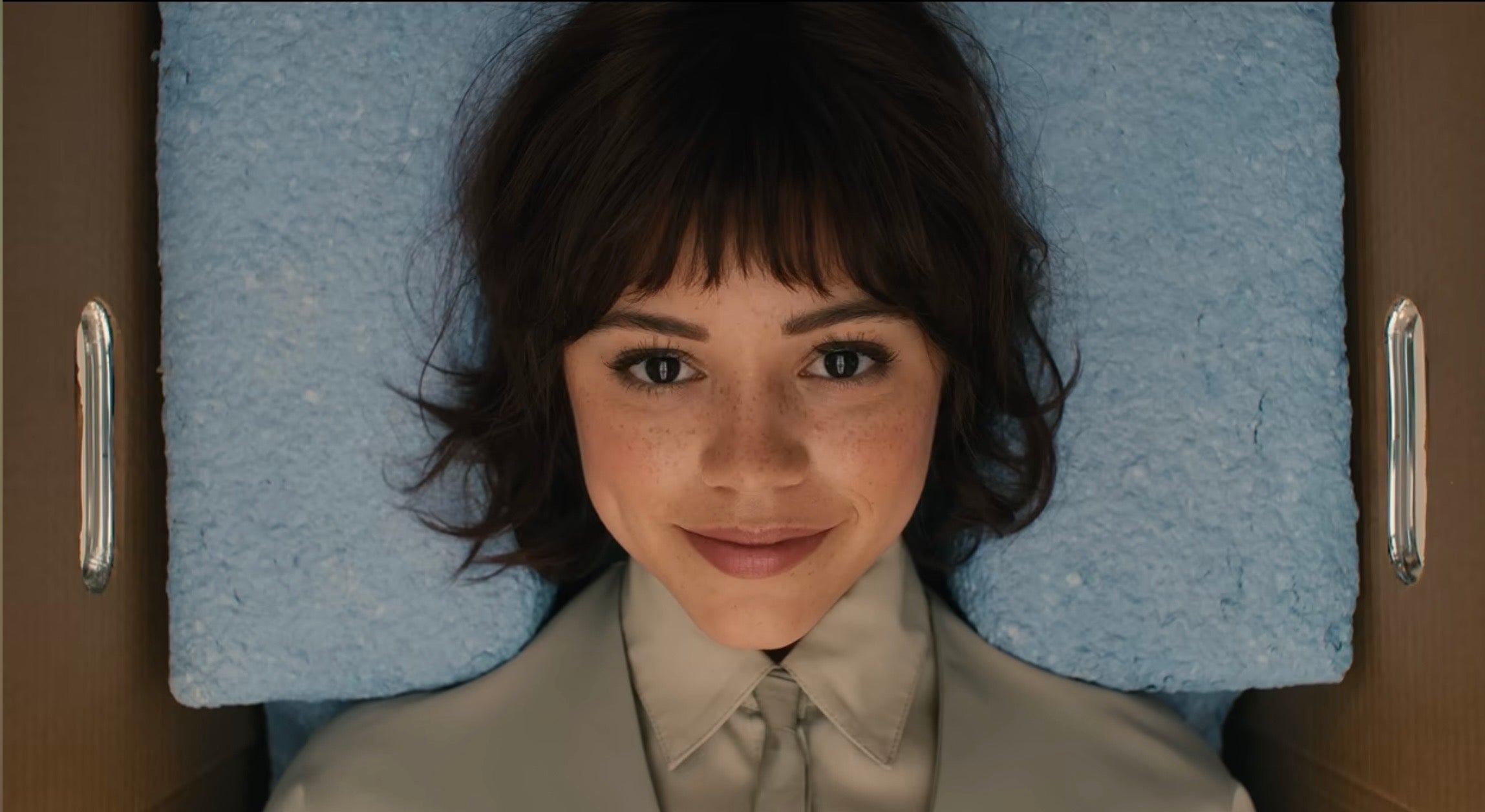 Jenna Ortega with short hair smiles while lying on a textured surface, wearing a collared shirt
