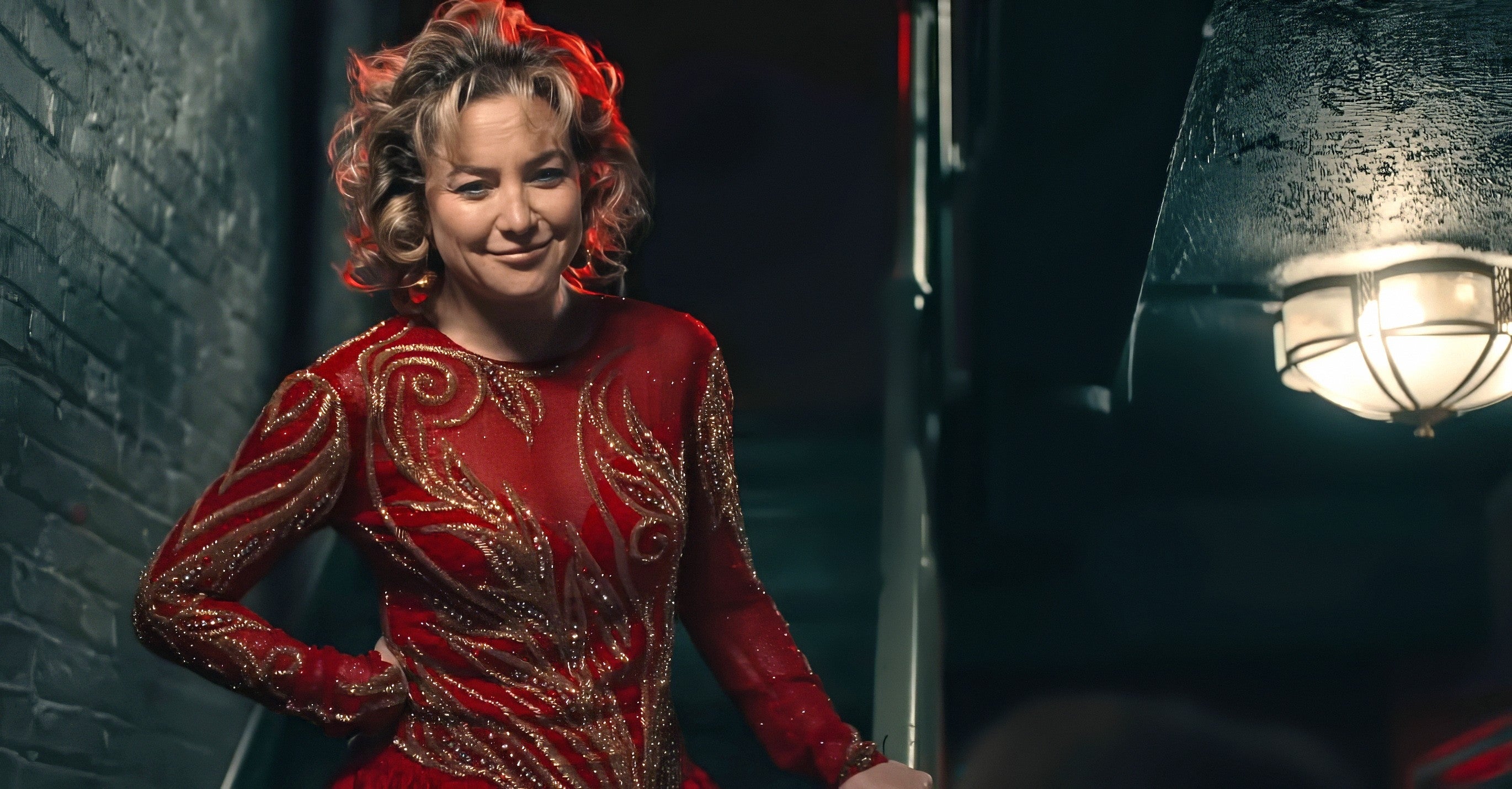 Kate Hudson stands confidently in ornate, patterned outfit on stairs indoors.