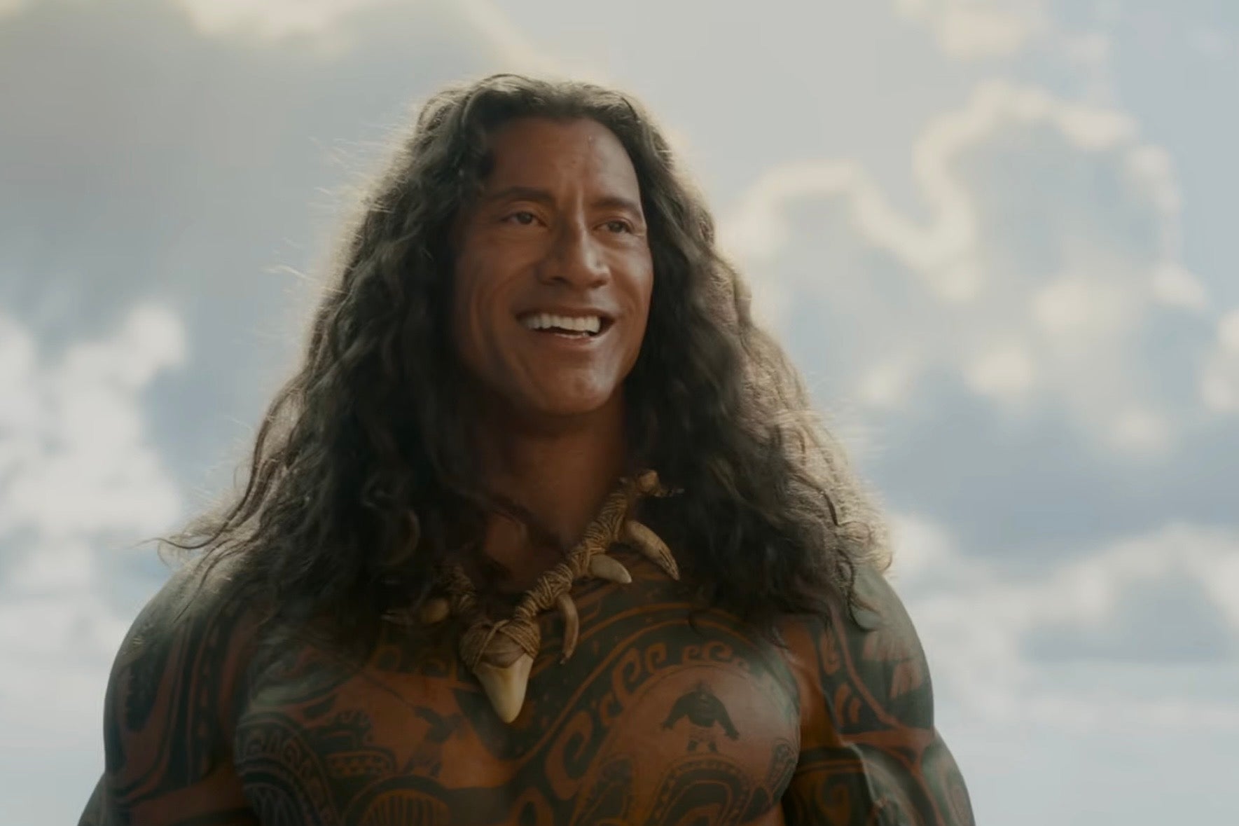 Dwayne Johnson with long hair and tribal tattoos stands smiling against a cloudy sky