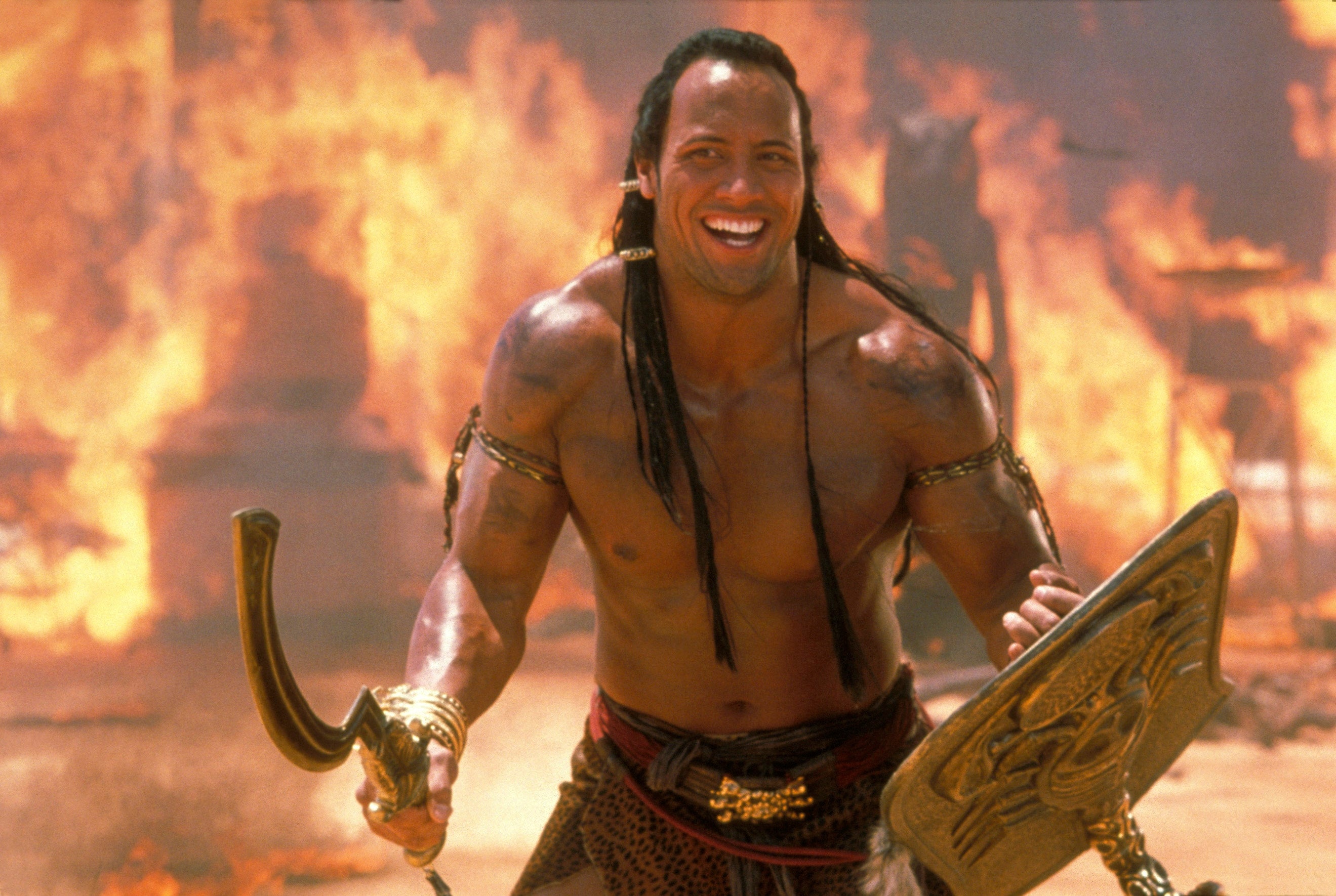 Dwayne Johnson with long hair holds a curved sword and shield, standing confidently amid flames