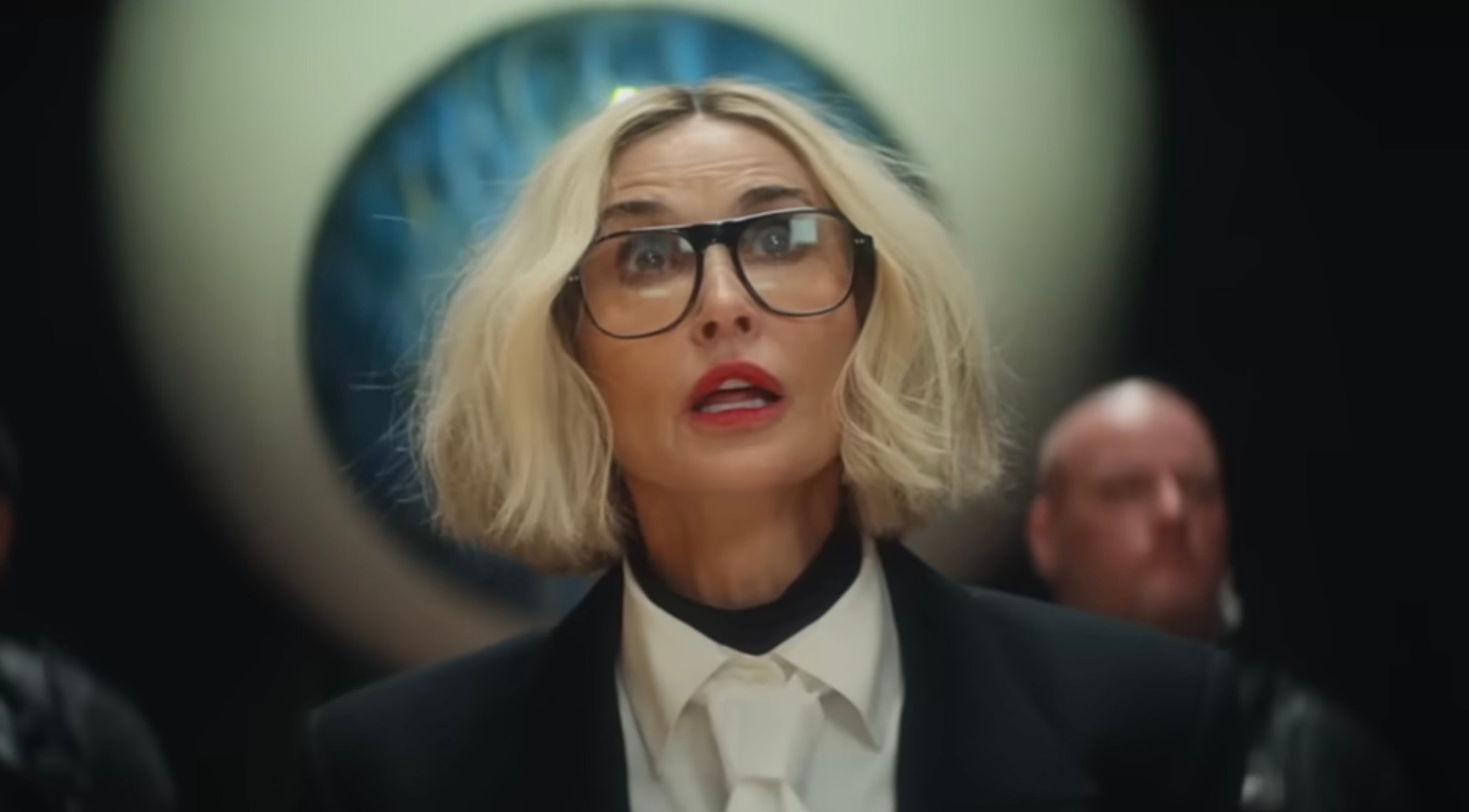 Demi Moore with short hair and large glasses wearing a formal black suit and white tie, appearing surprised or focused, with people and art in the background