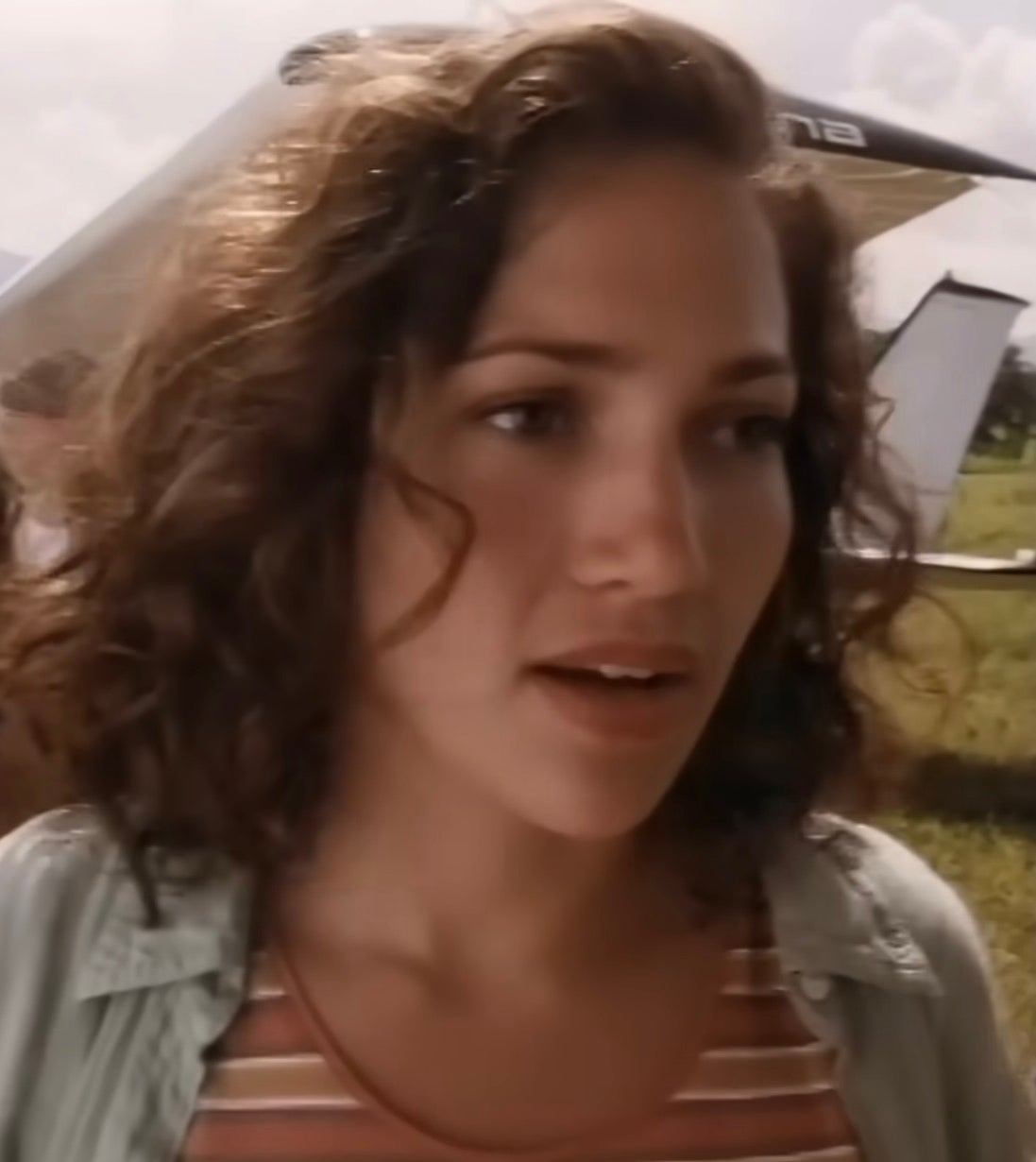 Jennifer Lopez with curly hair stands outdoors near an aircraft, wearing a striped shirt and open jacket, appearing thoughtful