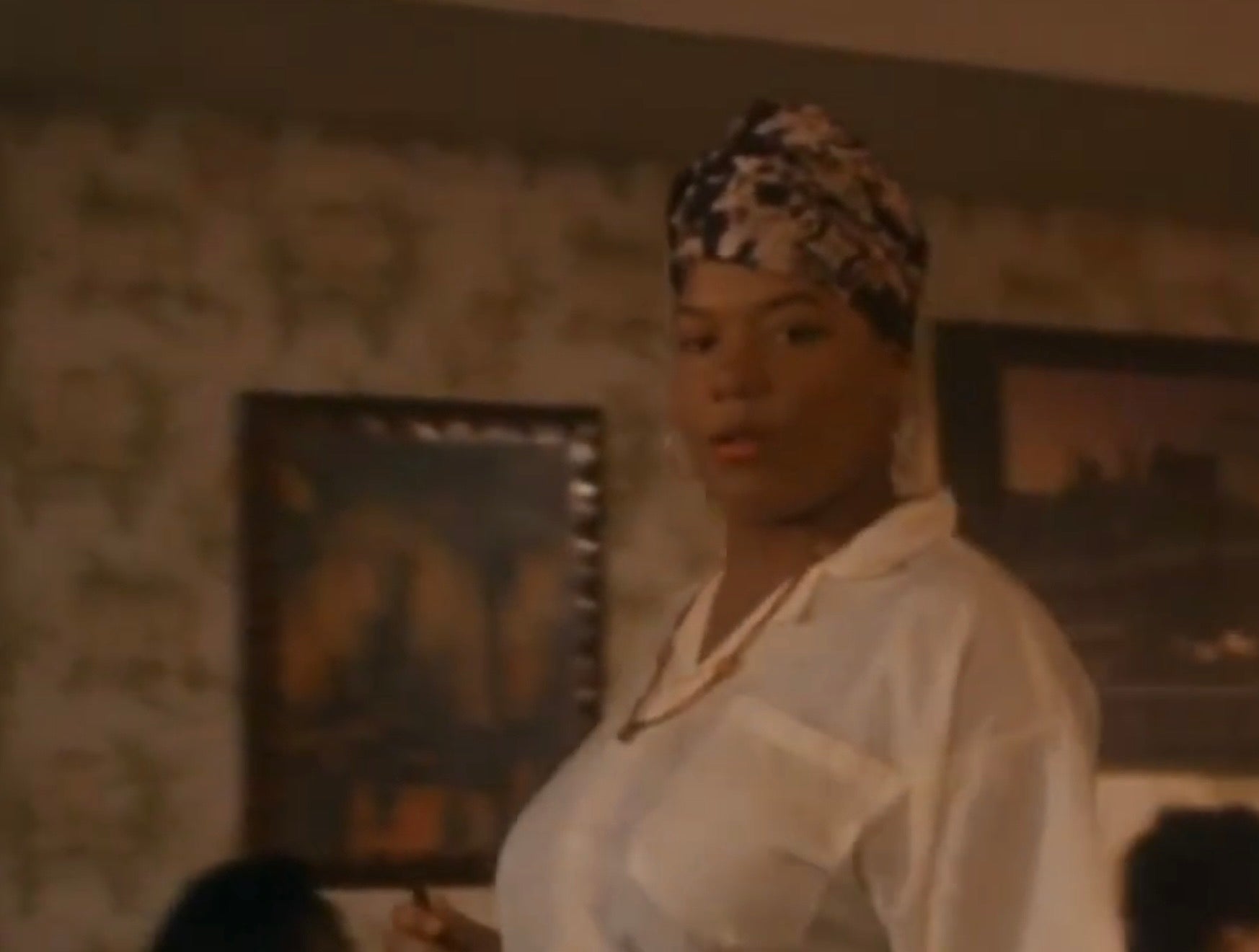 Younger Queen Latifah wearing a patterned headwrap and casual attire stands indoors with a confident expression