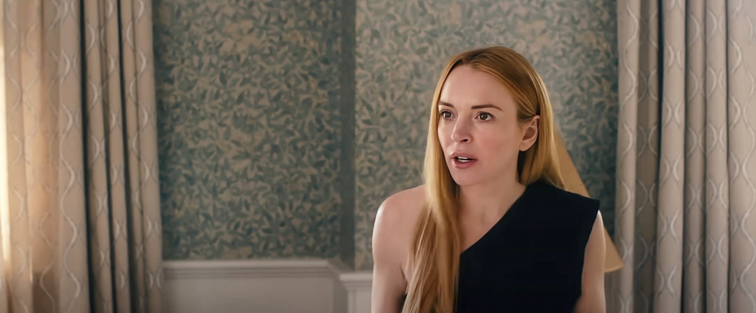 Lindsay Lohan with long hair looks surprised, wearing an asymmetrical top, standing indoors with patterned wallpaper in the background