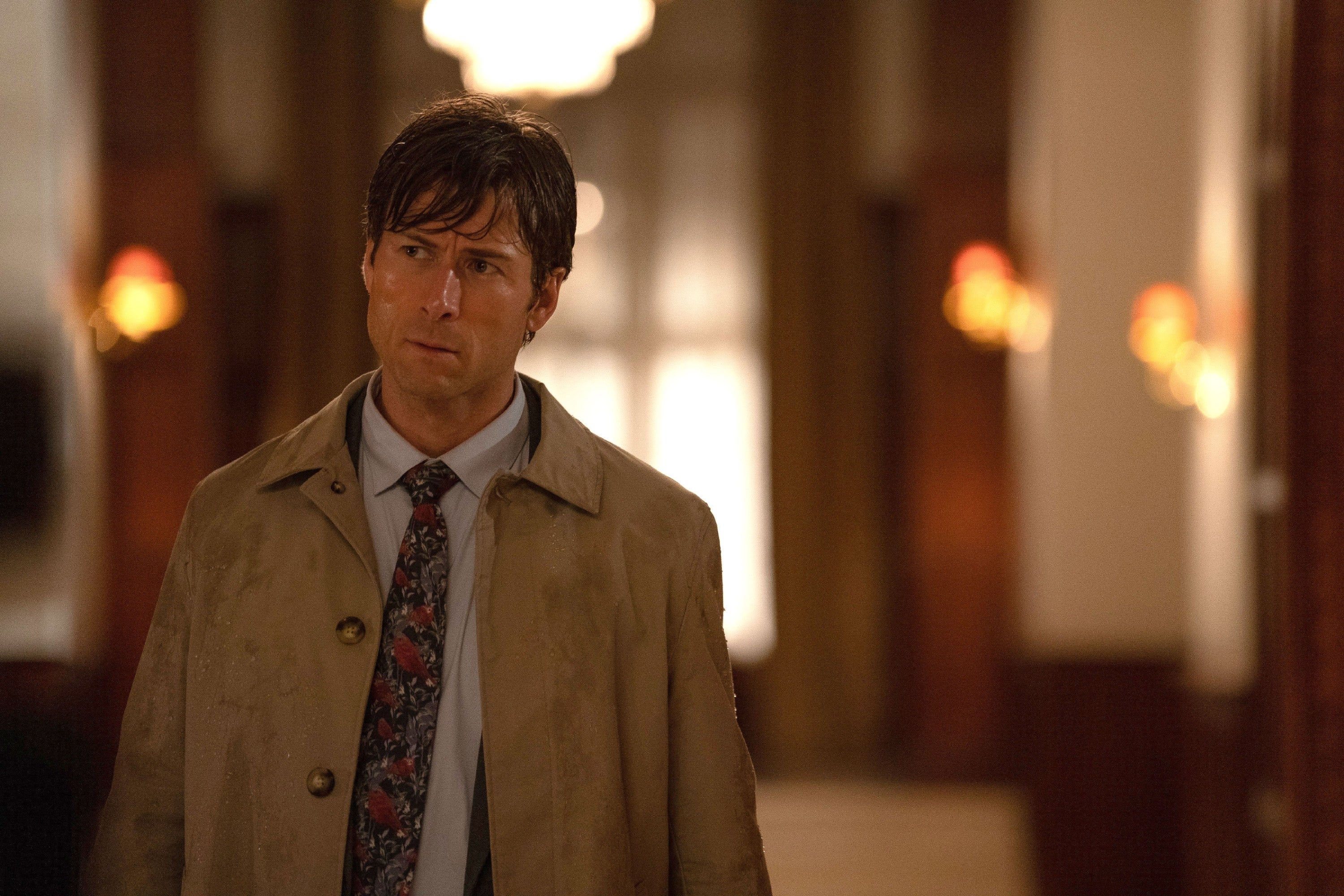 Glen Powell in a trench coat stands in a dimly lit hallway, looking pensive