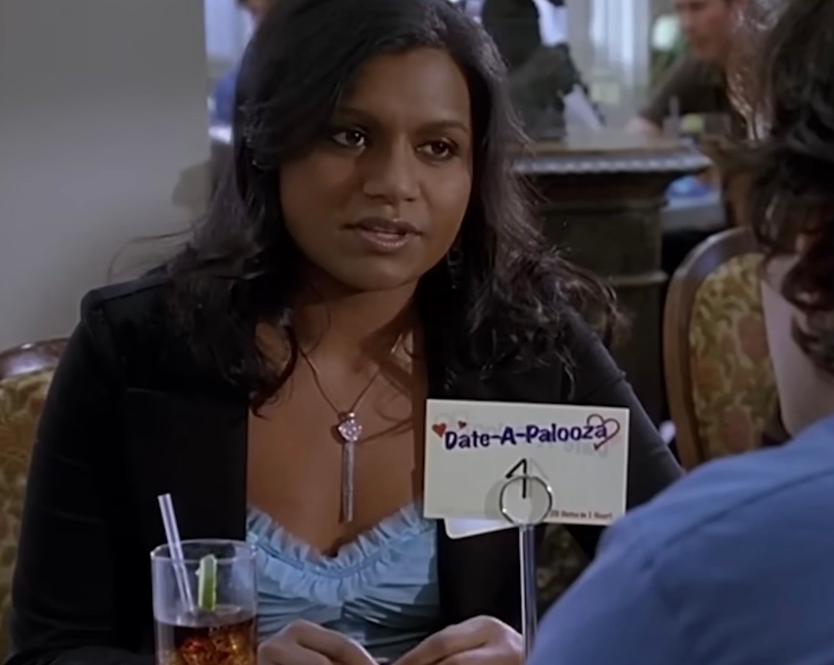 Younger Mindy Kaling at a speed dating event wearing a stylish blazer over a ruffled top, sitting at a table with a "Date-A-Palooza" sign