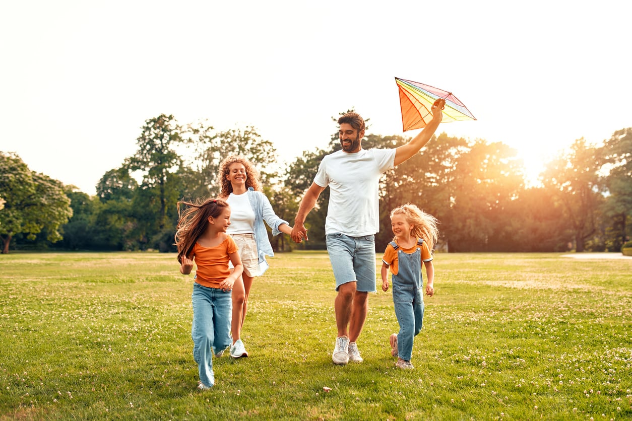 A family of four runs on grass at sunset, smiling. The father holds a colorful kite, while the mother and two daughters run beside him. Trees and sunlight are visible in the background.