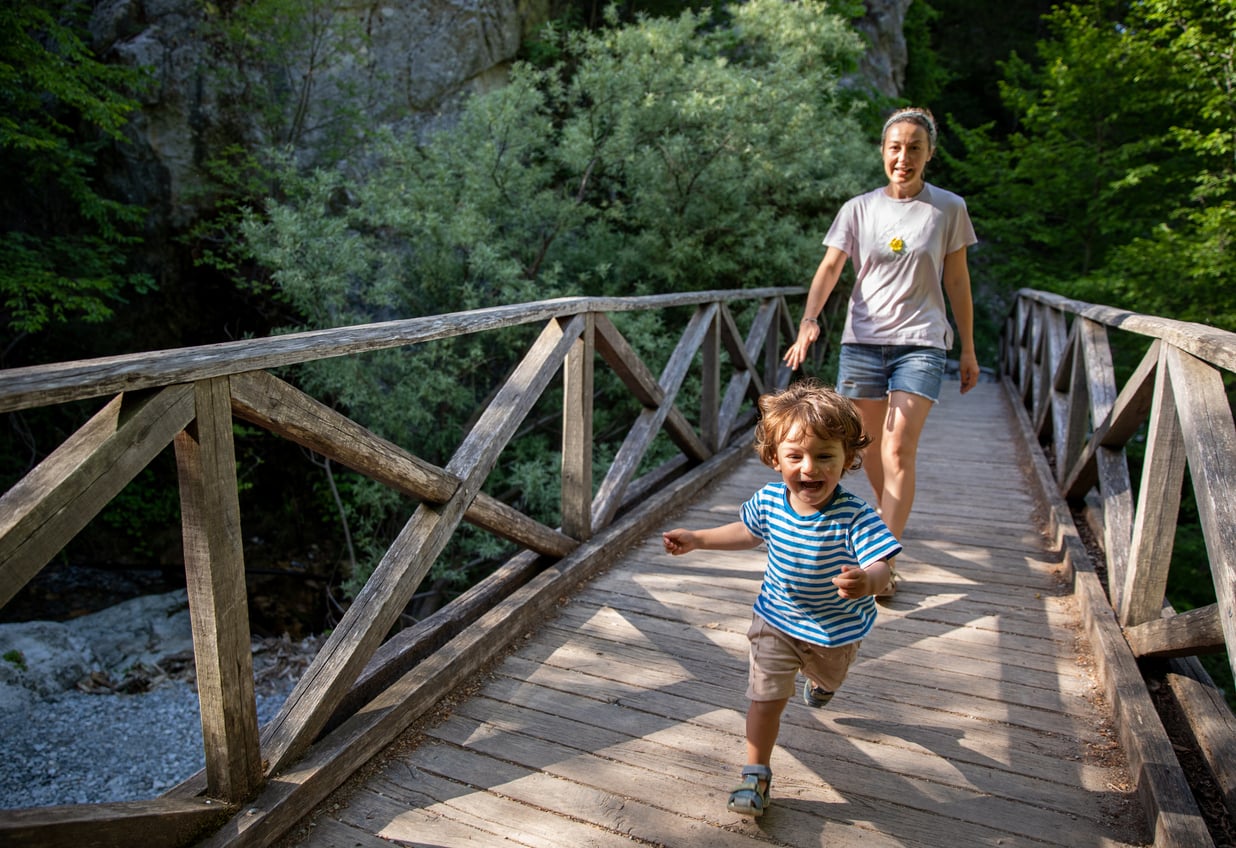 A young child runs joyfully across a wooden bridge, followed by an adult smiling behind. The scene is outdoors with lush green trees and sunlight filtering through.