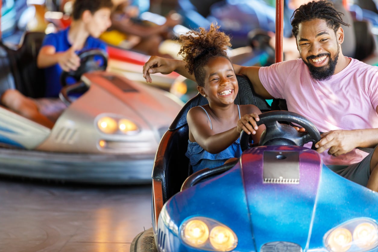 A smiling man and a young girl ride together in a blue bumper car at an amusement park, laughing and having fun. Other people in bumper cars are visible in the background.