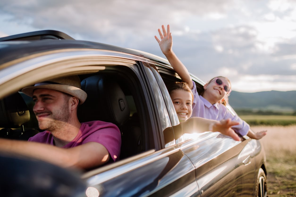 A man drives a car while two cheerful children lean out the windows, smiling and reaching their arms out. The car is in a scenic, sunny countryside area with hills and grass.