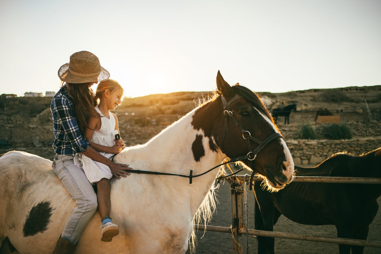 An adult and a child sit together on a horse at sunset, both smiling. The adult wears a straw hat and plaid shirt, while the child wears a white dress. They ride in a rustic outdoor setting with another horse nearby.