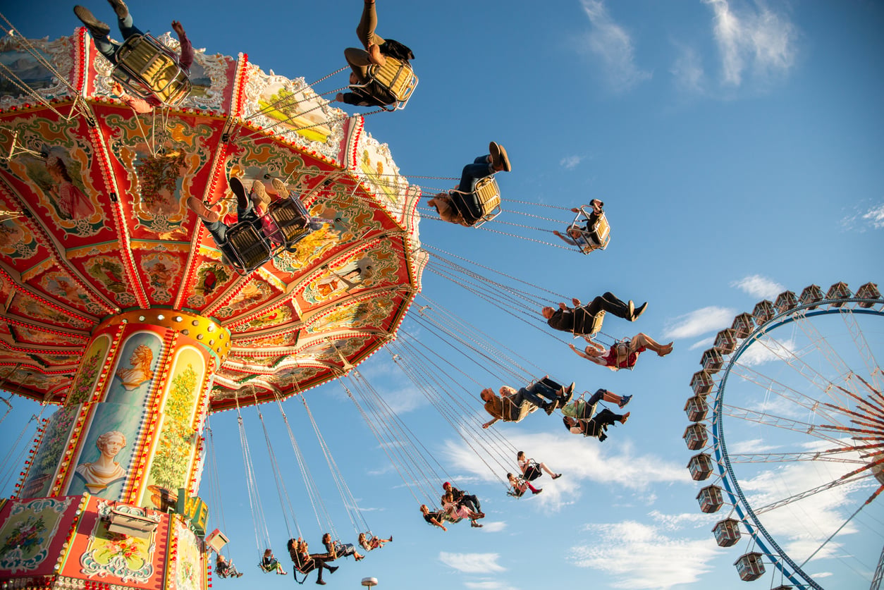People ride a colorful swing carousel at an amusement park. The ride spins as riders soar through the air, with a Ferris wheel and blue sky with clouds in the background.