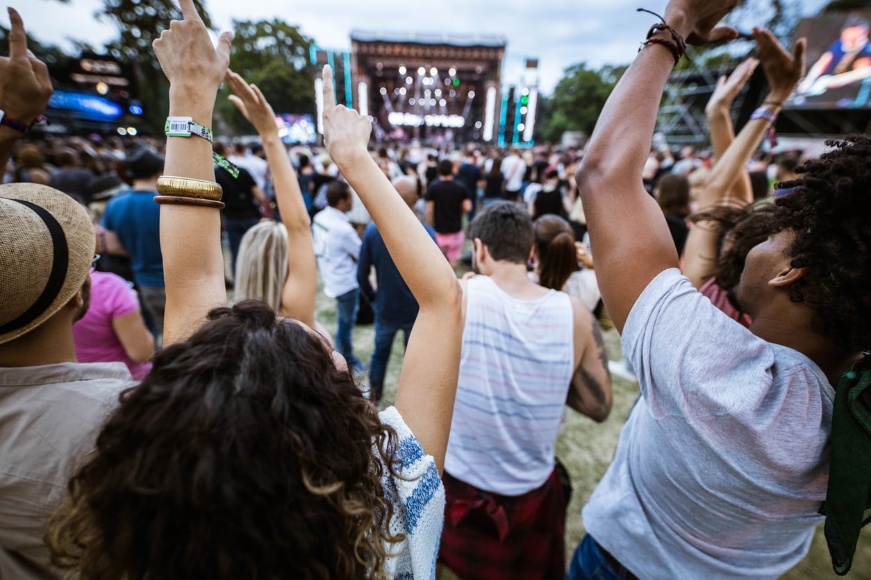 Crowd at an outdoor concert, people with raised arms facing a brightly lit stage in the distance. Trees and equipment surround the area, creating a lively festival atmosphere.