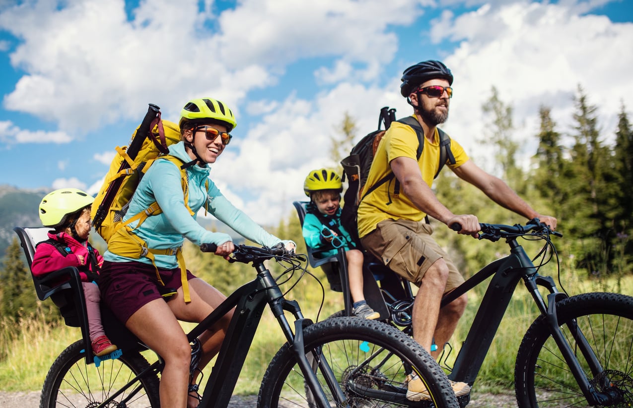 A family of four rides bicycles on a trail, with two kids in child seats. They all wear helmets and sunglasses, surrounded by greenery and blue sky with clouds.