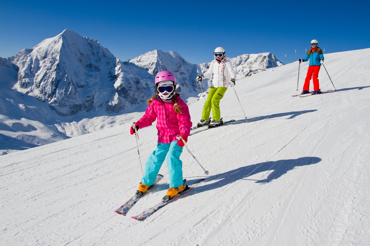 Three people skiing down a snowy mountain slope on a sunny day, with tall, snow-covered peaks visible in the background. The front skier is a child in bright clothes, followed by two adults.