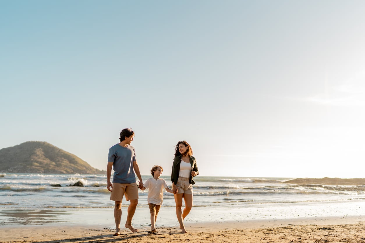 A family of three walks hand in hand along a sandy beach with the ocean and a hill in the background under a clear sky.