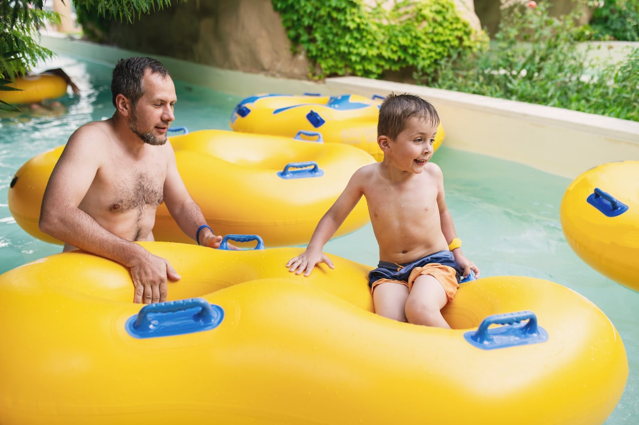 A man and a young boy sit on large yellow inflatable tubes in a lazy river at a water park, surrounded by greenery.