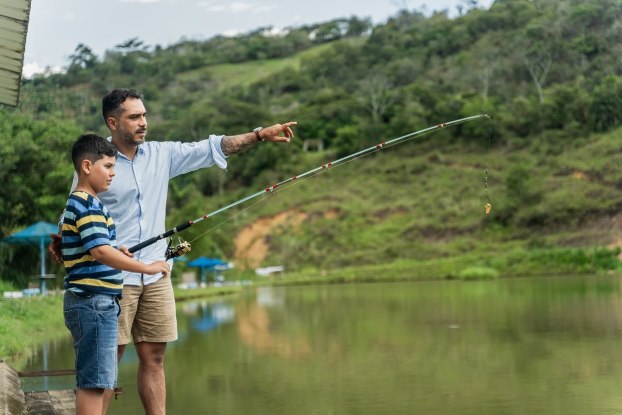 A man and a boy stand by a lake; the man points ahead while the boy holds a fishing rod. Green hills and trees are in the background.