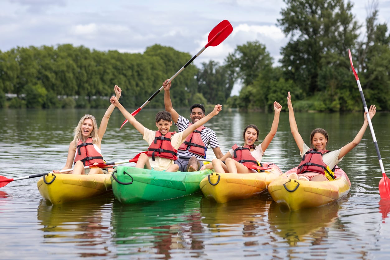 Four people in two yellow kayaks and one green kayak, all wearing life jackets, smiling and raising their arms in celebration on a calm lake, with trees and cloudy sky in the background.