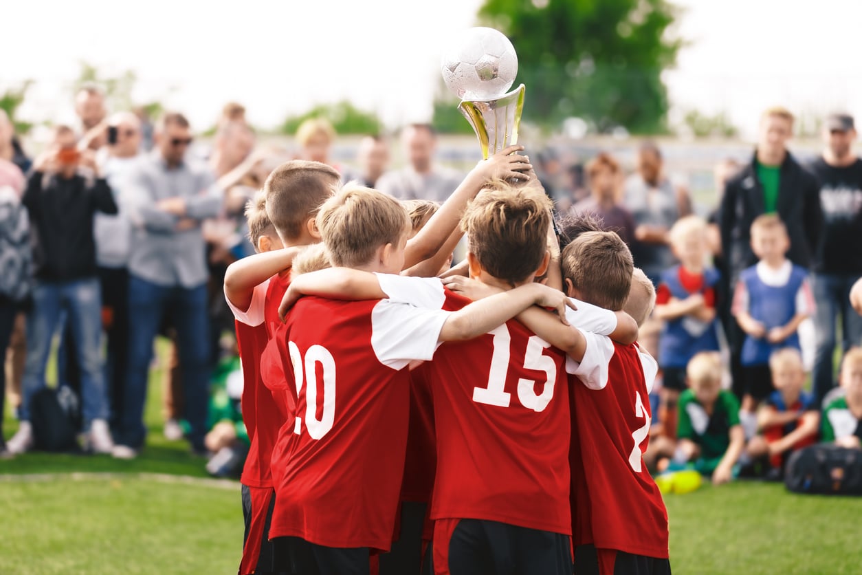 A youth soccer team in red jerseys celebrates together, holding up a trophy on a grassy field while a crowd of spectators watches and cheers in the background.
