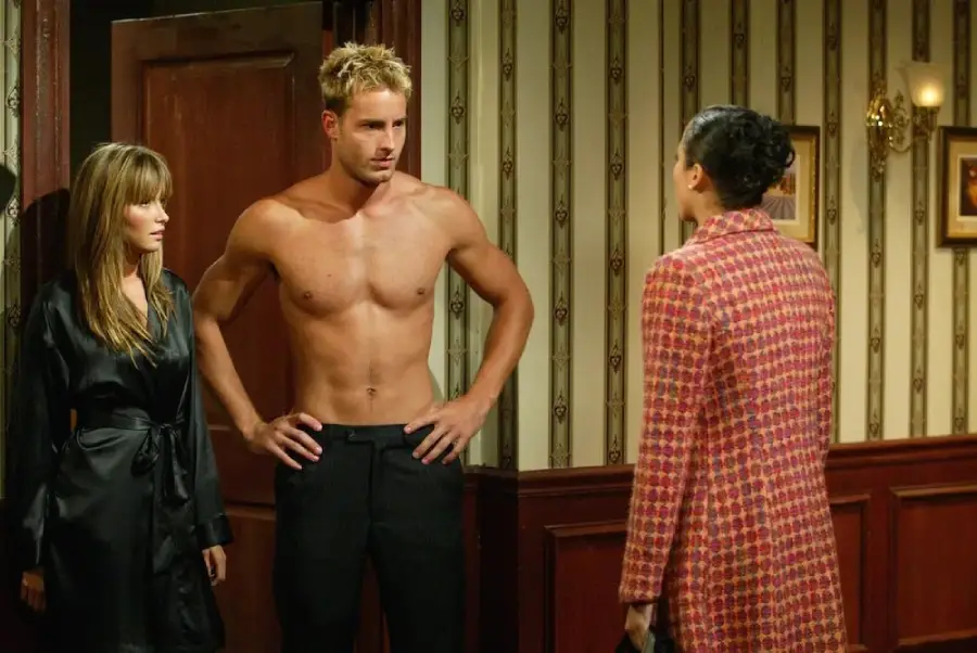 A shirtless man stands between a woman in a black robe and another woman in a red patterned coat, all inside a room with striped wallpaper and framed pictures.