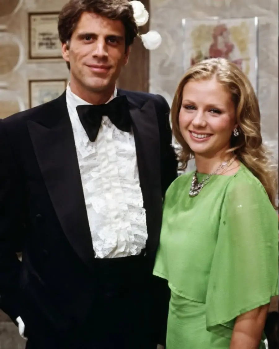 A man in a black tuxedo with a ruffled white shirt stands next to a woman in a light green dress. Both are smiling and posing indoors with framed artwork visible in the background.