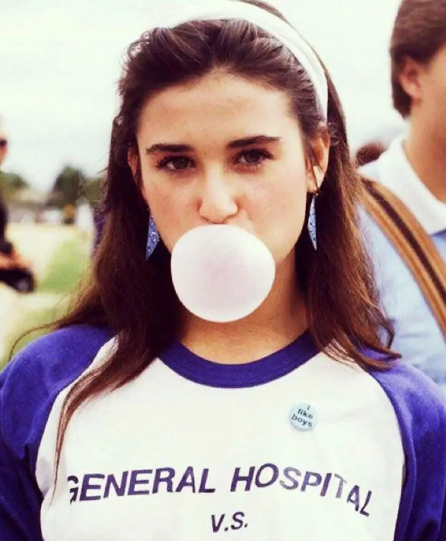 Young woman with long dark hair blows a bubblegum bubble. She wears a blue and white "General Hospital V.S." shirt and a headband. The background is outdoors and slightly blurred.